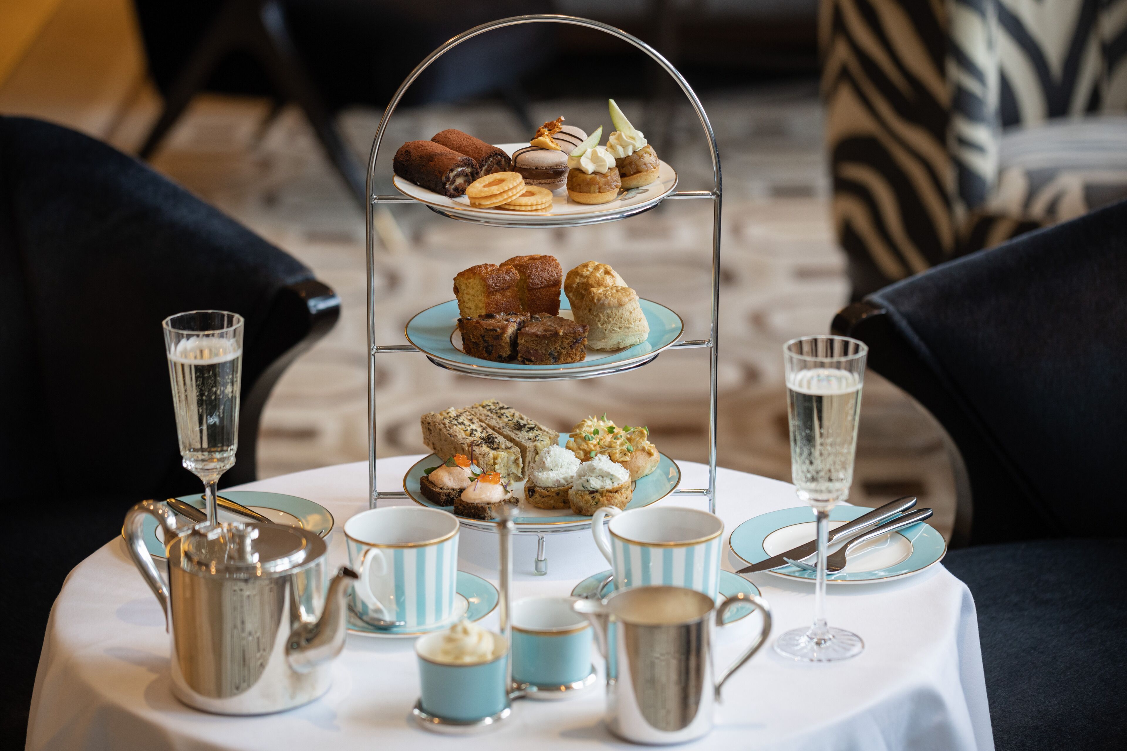 A three-tiered dessert stand with various pastries and a glass of champagne on a table, surrounded by matching cups and a silver teapot.