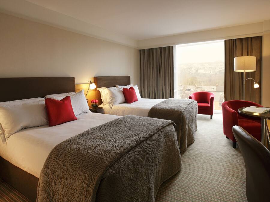 Quarto de hotel com duas camas, uma escrivaninha, uma cadeira e vista para a cidade.
