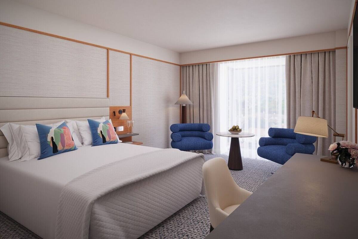 Une chambre d’hôtel avec un grand lit, deux fauteuils bleus, une petite table ronde et un bureau avec une lampe.