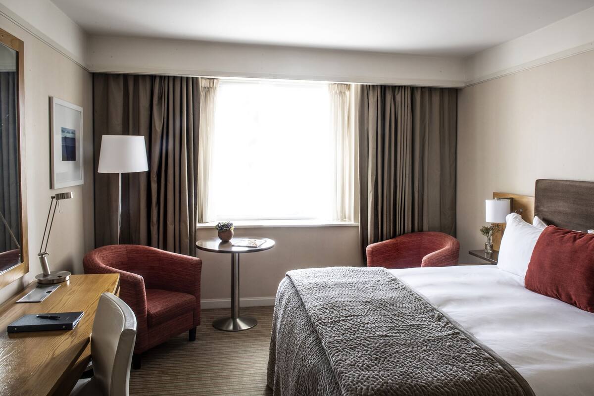 Une chambre d’hôtel avec un lit, deux fauteuils, une petite table, une lampe et une fenêtre avec des rideaux.