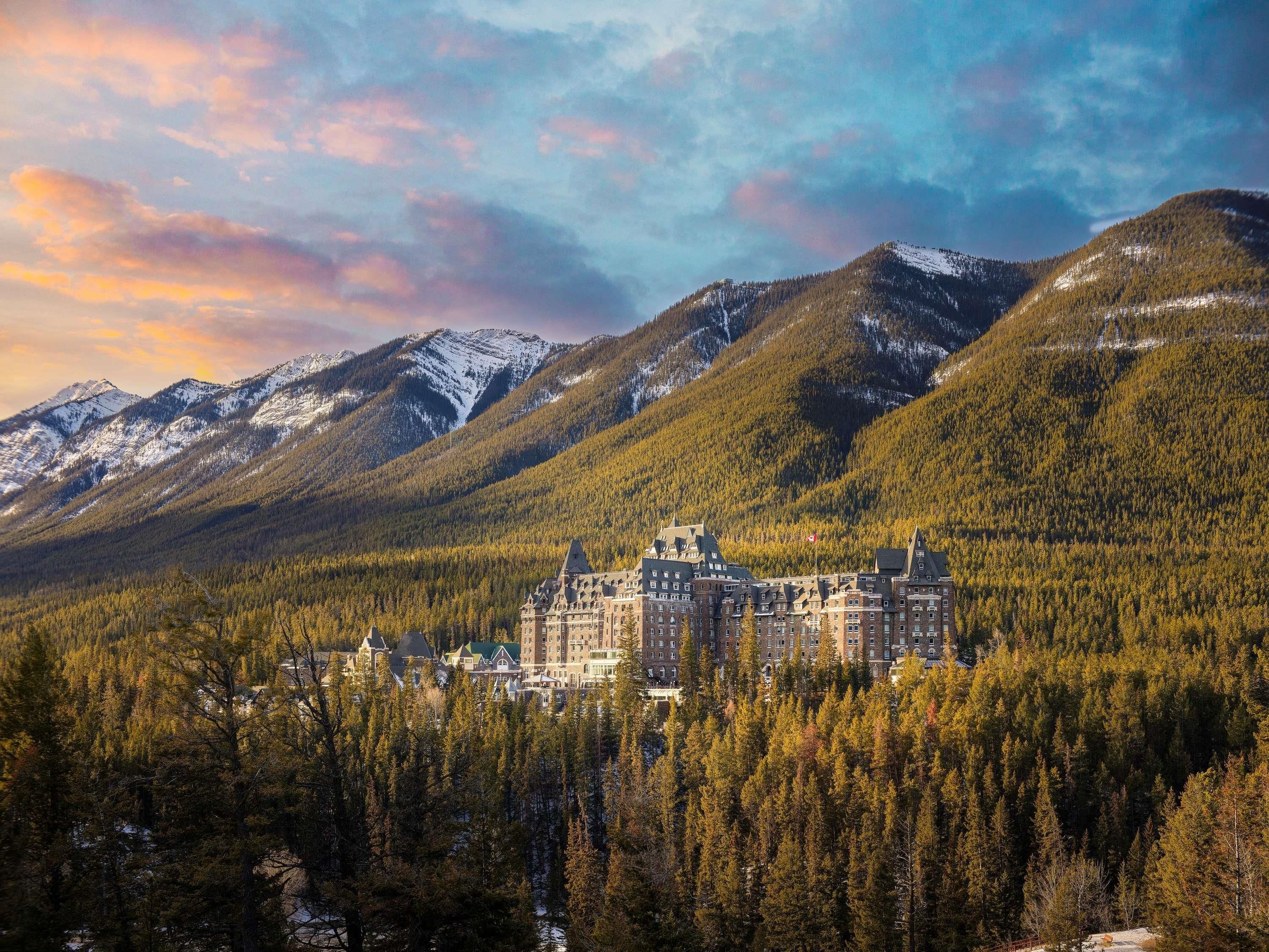 A historic hotel with a central building surrounded by forested hills and snow-capped mountains.
