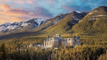 A historic hotel with a central building surrounded by forested hills and snow-capped mountains.