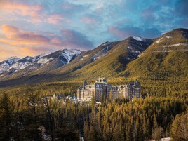 Un hotel histórico con un edificio central rodeado de colinas boscosas y montañas nevadas.