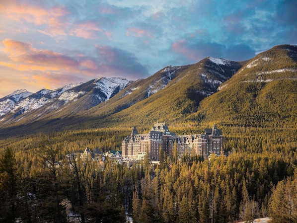 A historic hotel with a central building surrounded by forested hills and snow-capped mountains. - Fairmont Banff Springs (Banff)