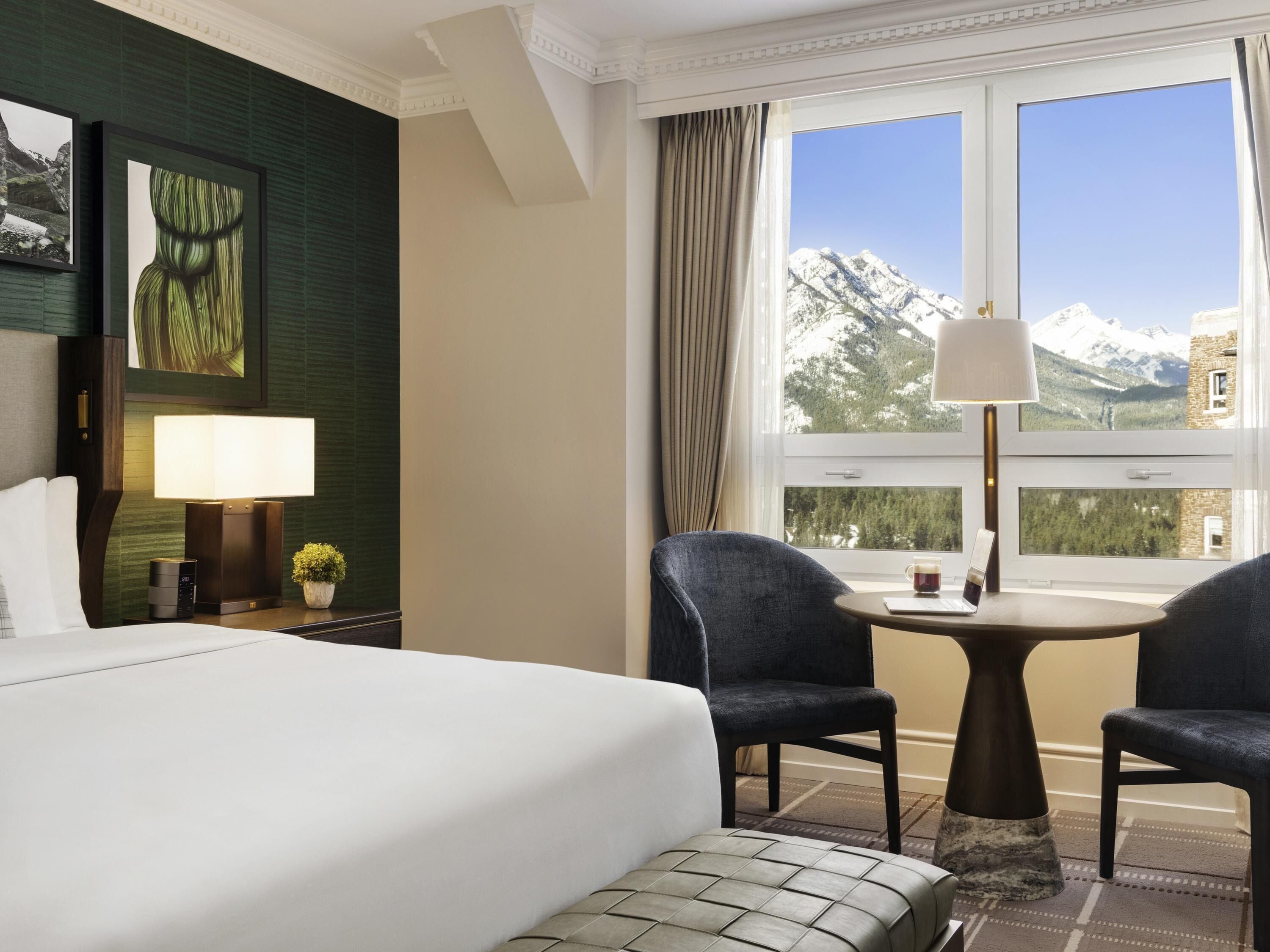 Une chambre d’hôtel avec un lit, deux chaises, une petite table, une lampe, une table de chevet et une fenêtre donnant sur la montagne.