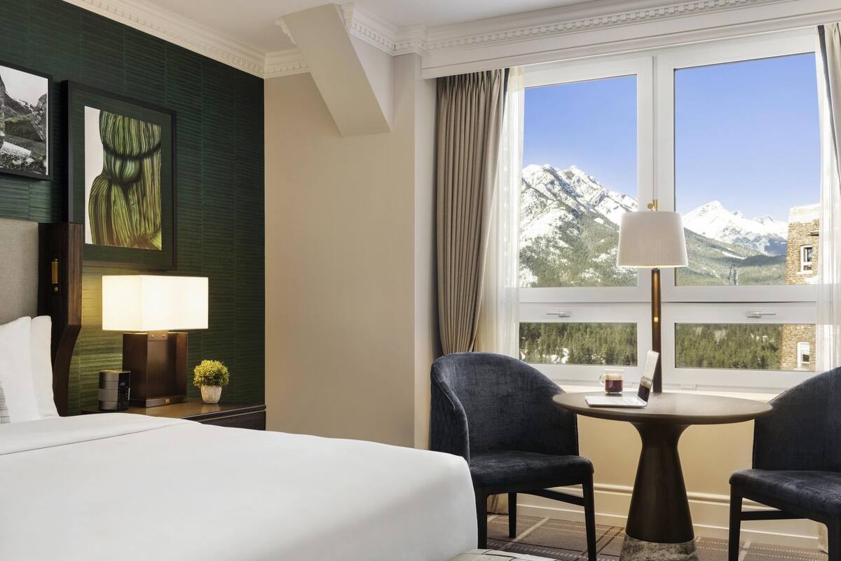 A hotel room with a bed, two chairs, a small table, a lamp, a nightstand, and a window with a mountain view.