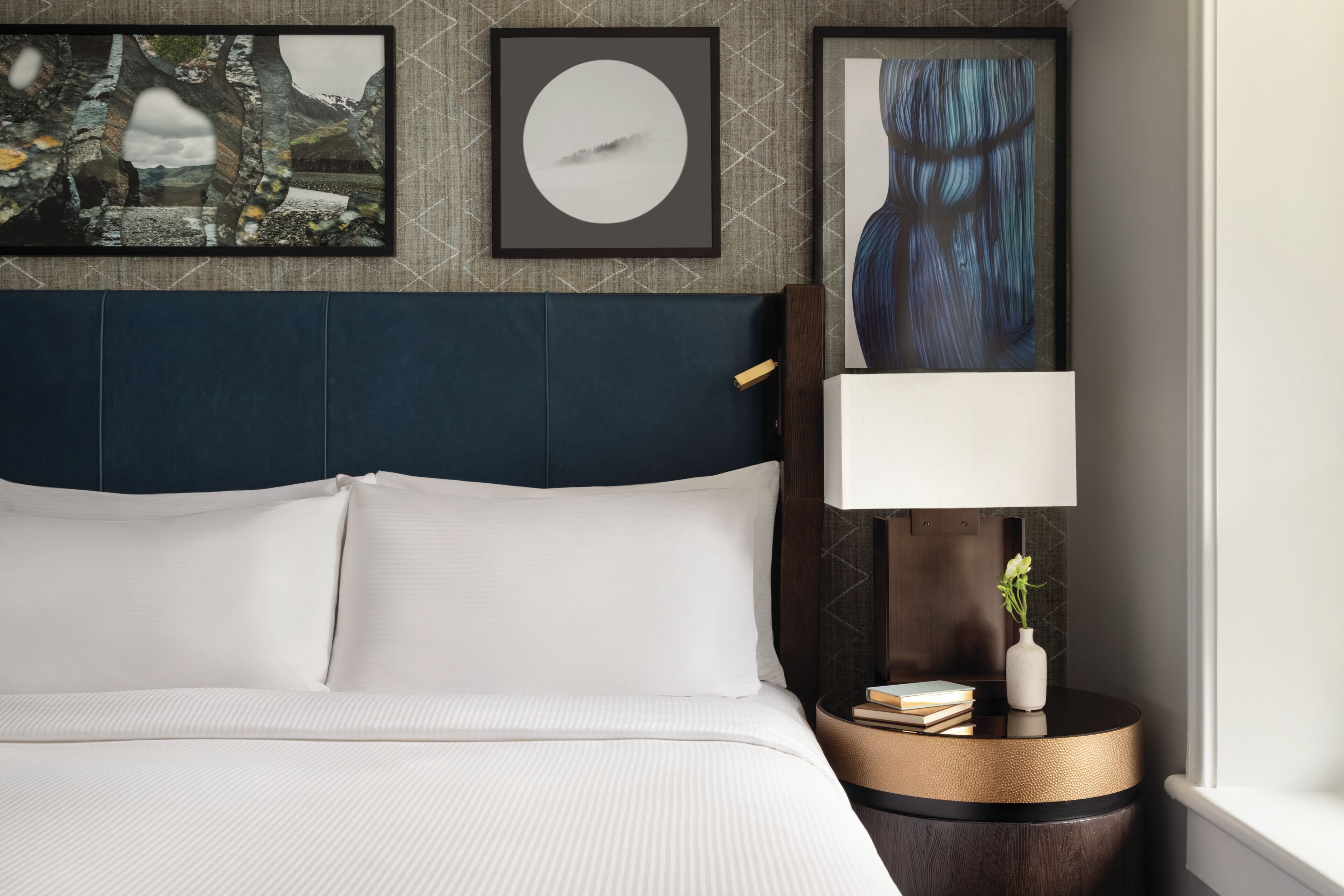A neatly made bed with white linens, a dark blue headboard, and a bedside table with a lamp, books, and a vase.