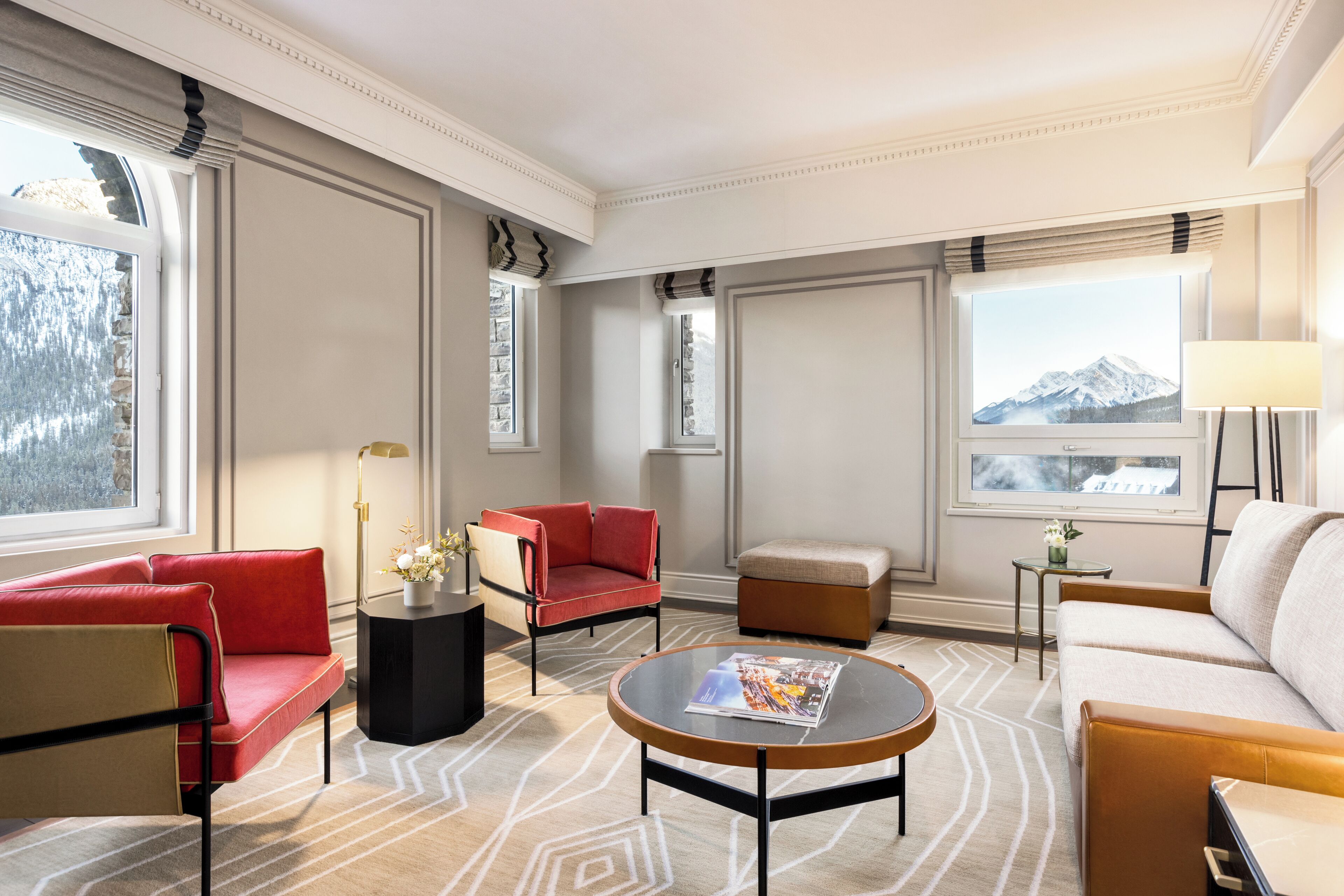 Una habitación de hotel moderna con un sofá rojo, un sillón color beige, una mesa de centro redonda y vistas a la montaña a través de amplias ventanas.