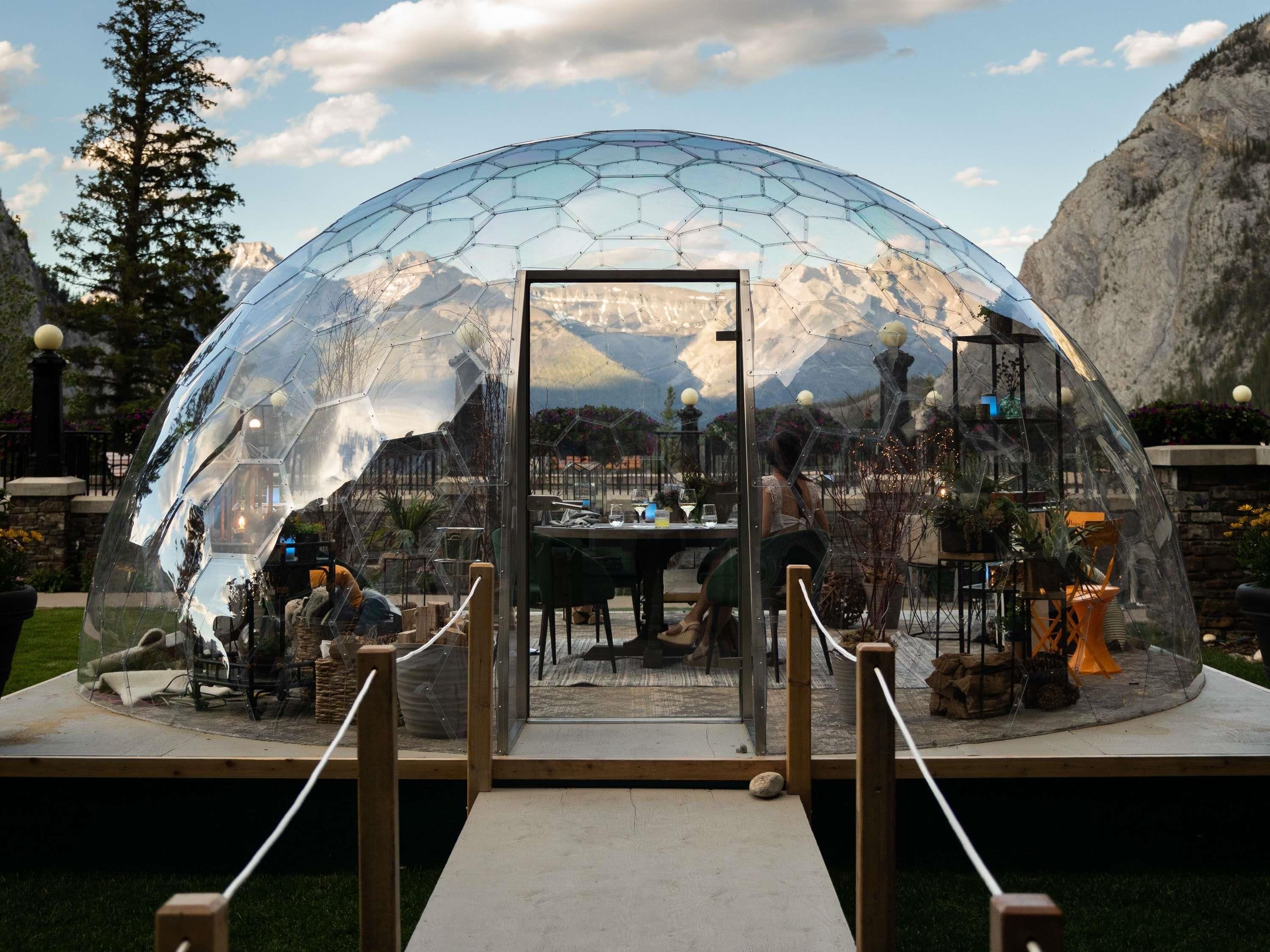 A transparent dome structure with a wooden platform and railing in front, surrounded by a mountainous landscape.