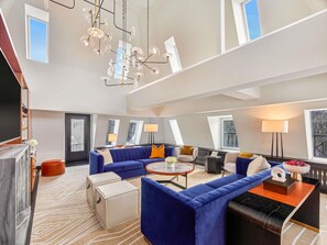 A spacious living room with a high ceiling, a modern chandelier, and a large window providing natural light.