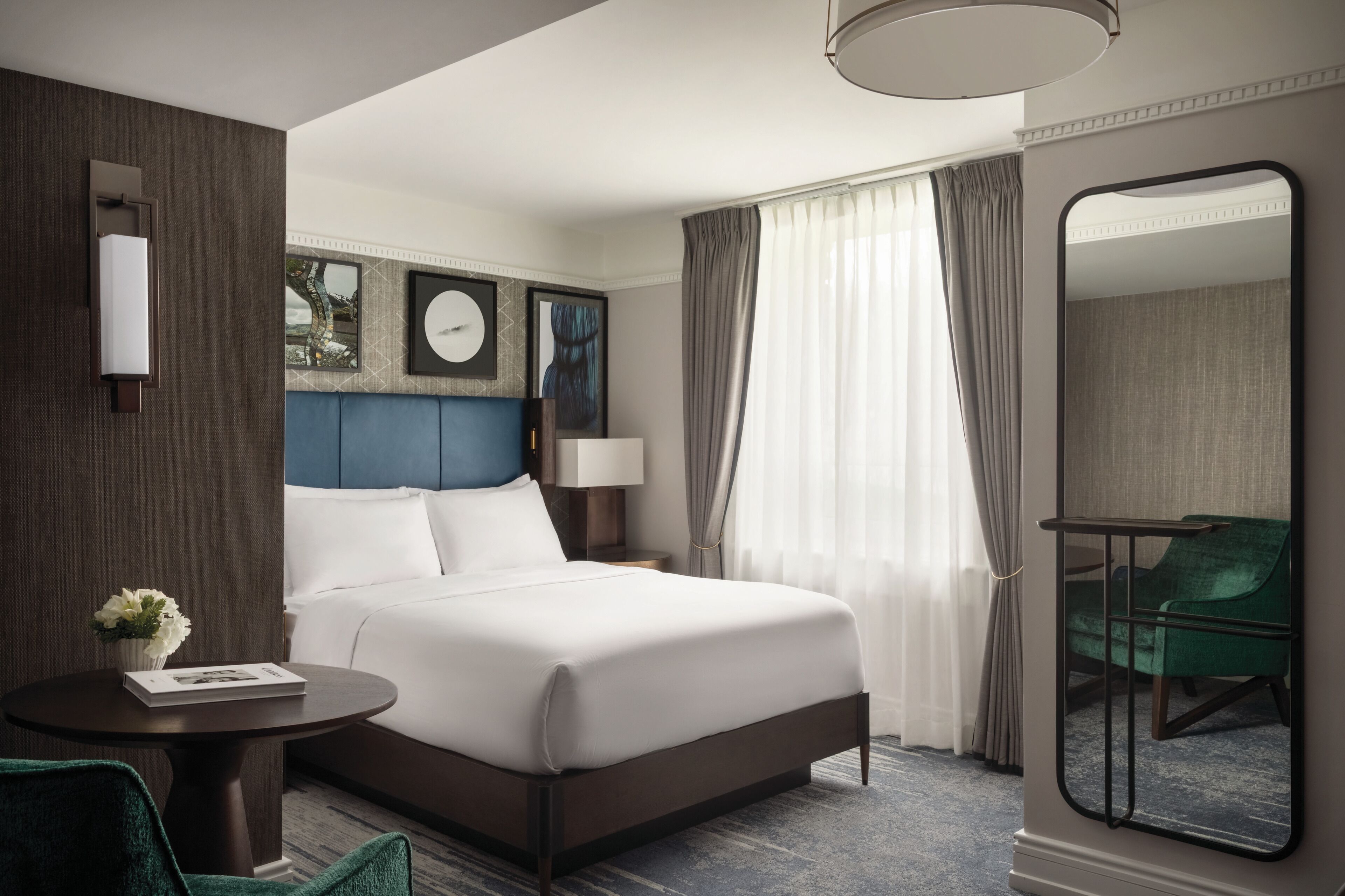 A hotel room with a large bed, a round table with a vase of flowers, a green chair, a mirror, and a view of the city through the window.