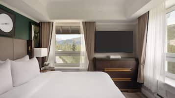 A hotel room with a large bed, a desk with a flat-screen TV, a window with curtains, and a view of mountains.