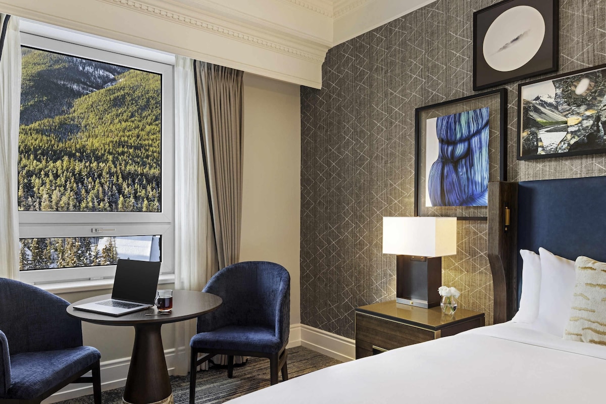 A hotel room with a bed, a desk with a laptop, two chairs, a nightstand with a lamp, and a large window with a view of a forest.