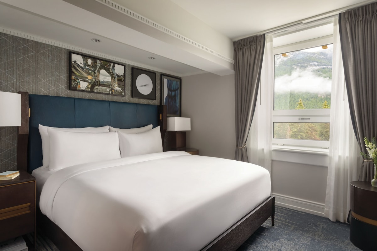A hotel room with a large bed, a nightstand, a lamp, a window with curtains, and artwork on the wall.