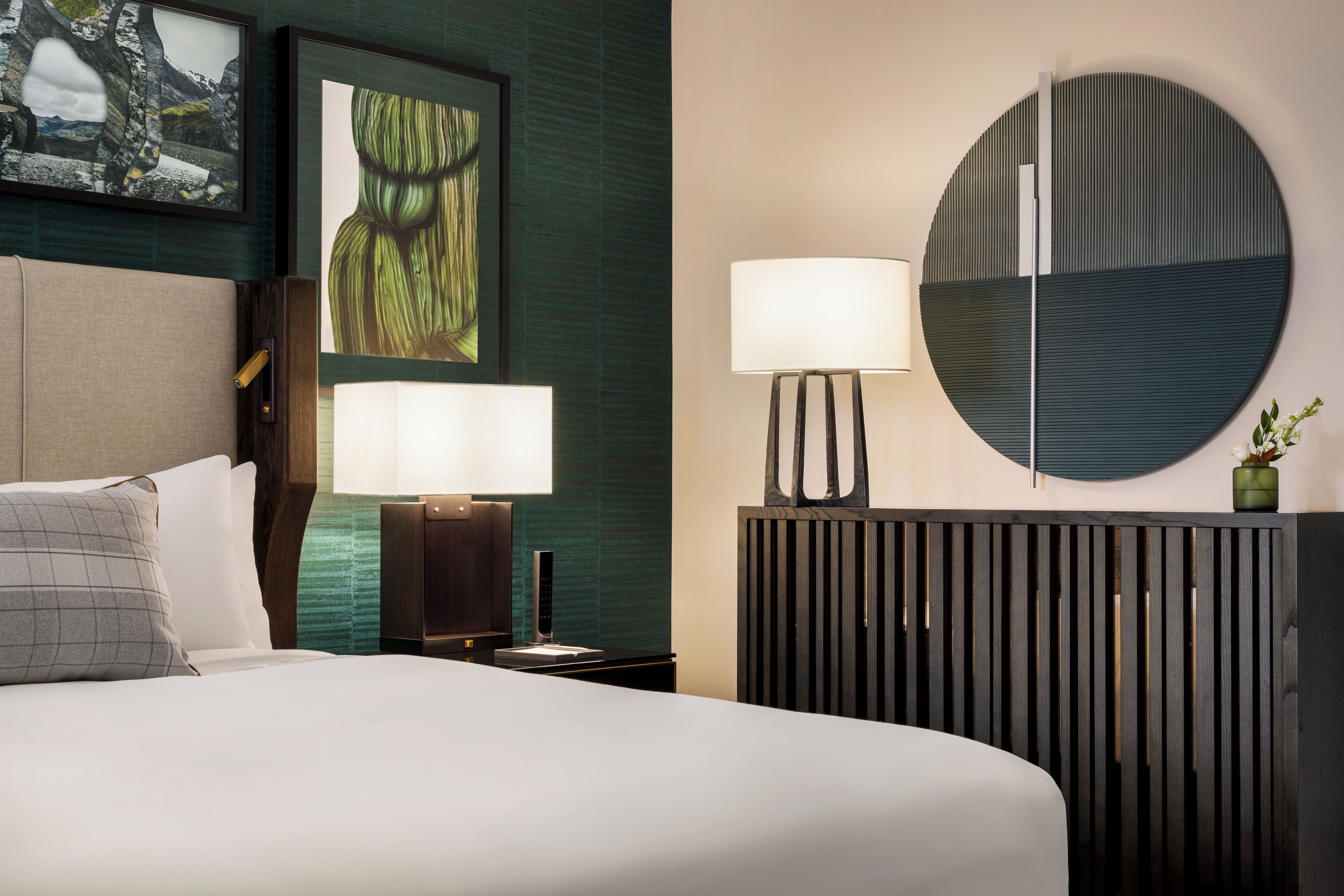 A modern hotel room with a bed, two bedside lamps, a round mirror, and a decorative plant.