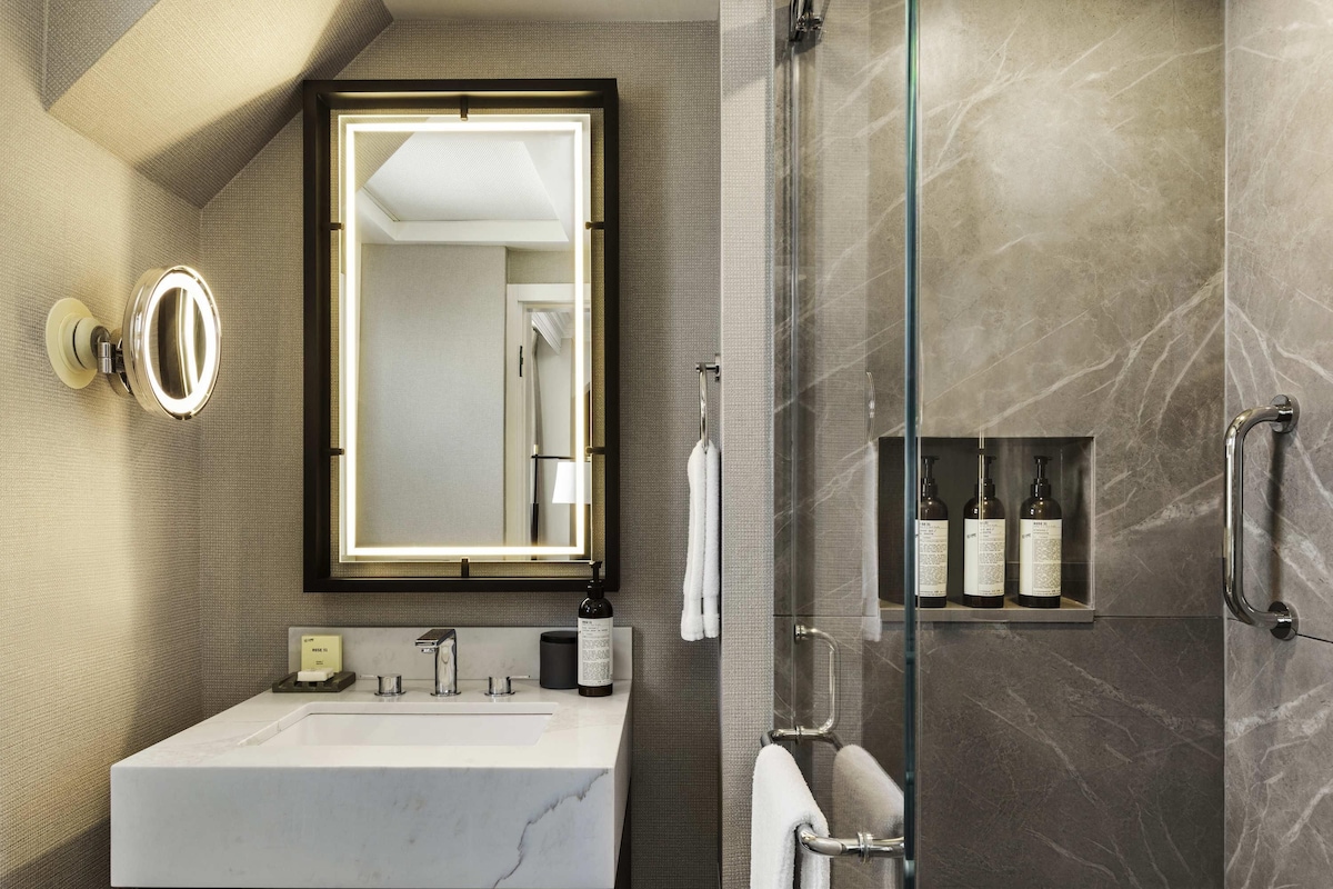 A modern bathroom with a marble sink, a large mirror with lights, a glass shower enclosure, and a shelf with toiletries.