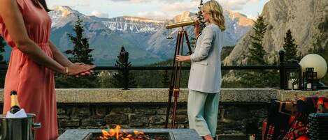 A terrace with a stone fireplace, a telescope, and a mountainous backdrop.
