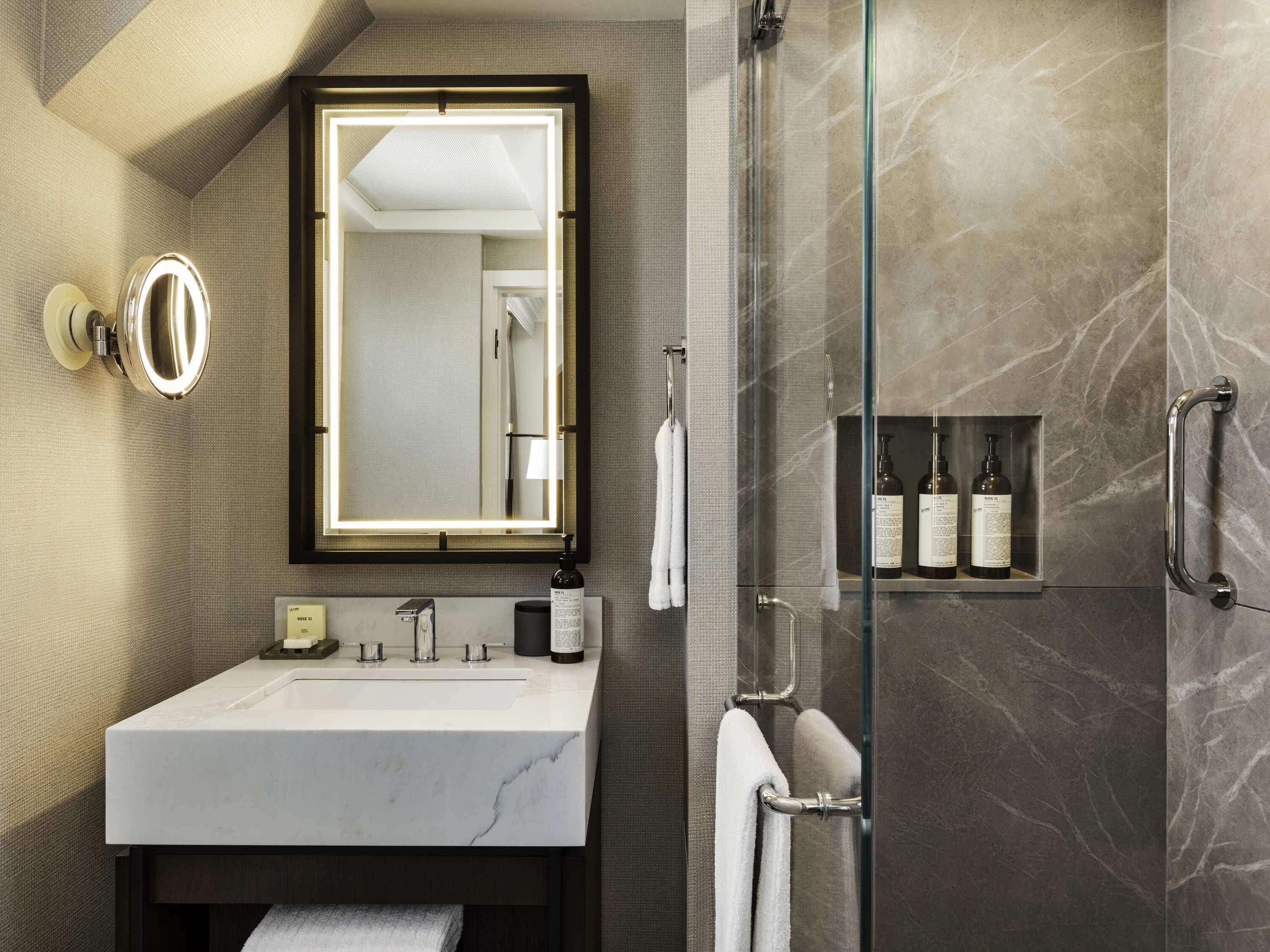 Une salle de bain moderne avec un lavabo en marbre, un grand miroir avec éclairage intégré, une douche en verre et une étagère contenant des articles de toilette.