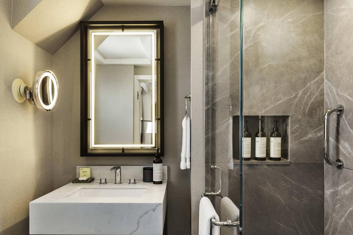A modern bathroom with a marble sink, a large mirror with lights, a glass shower enclosure, and a shelf with toiletries.