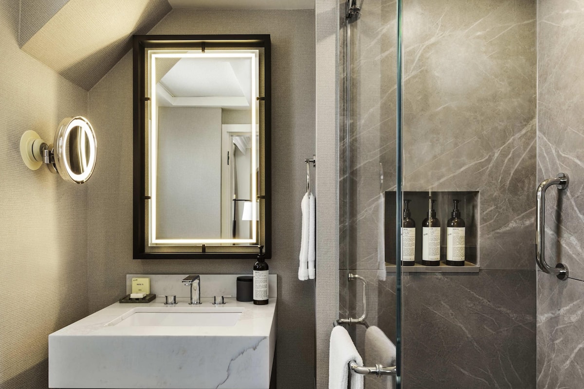 Une salle de bain moderne avec un lavabo en marbre, un grand miroir avec éclairage intégré, une douche en verre et une étagÚre contenant des articles de toilette.