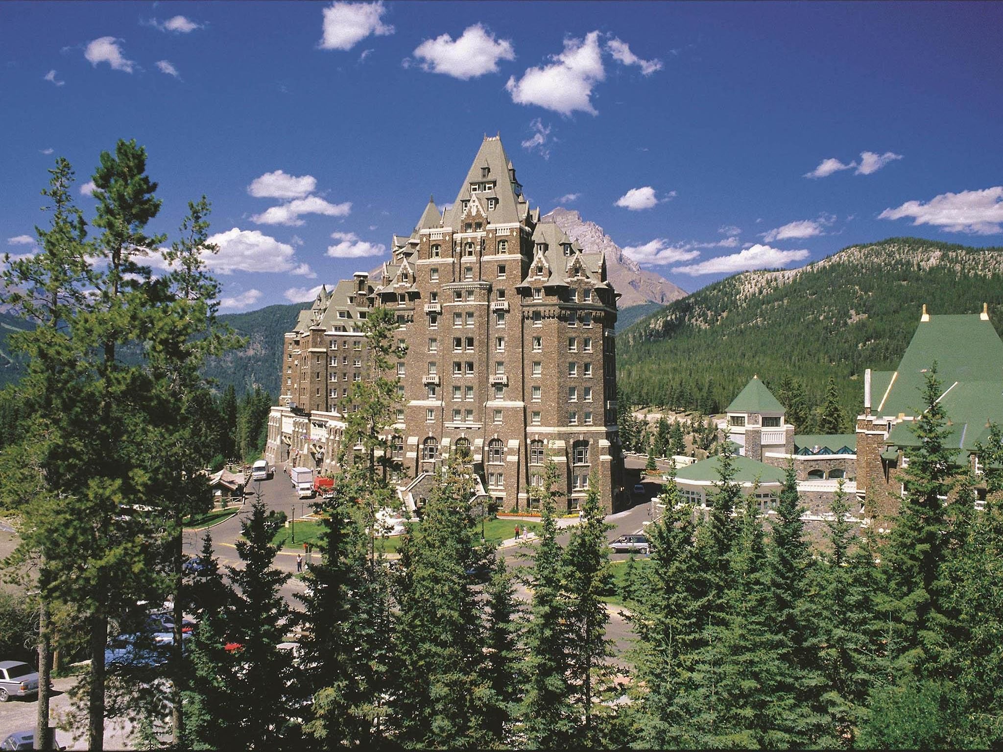 A large, historic hotel with a prominent tower, surrounded by dense forest and mountains.