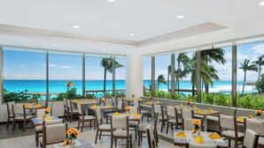 Restaurante com mesas e cadeiras dispostas para refeições, grandes janelas oferecendo vista para o oceano e palmeiras.