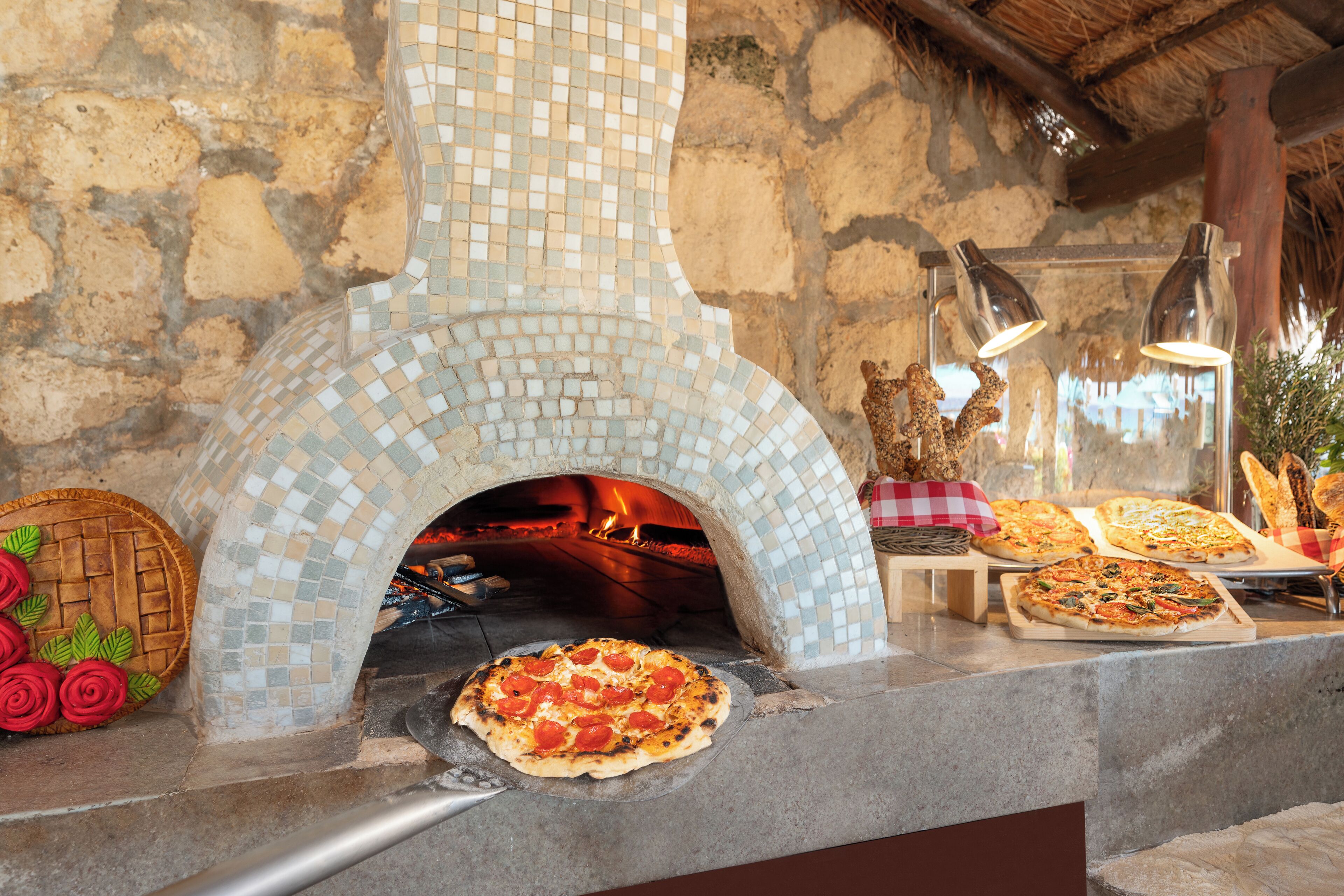 Een stenen muur met een oven betegeld met mozaïek, pizza op een pizzaschep, en verschillende pizza's op een toonbank.
