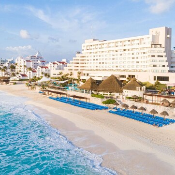 A beachfront hotel with multiple floors, a beach with blue chairs, and thatched umbrellas.