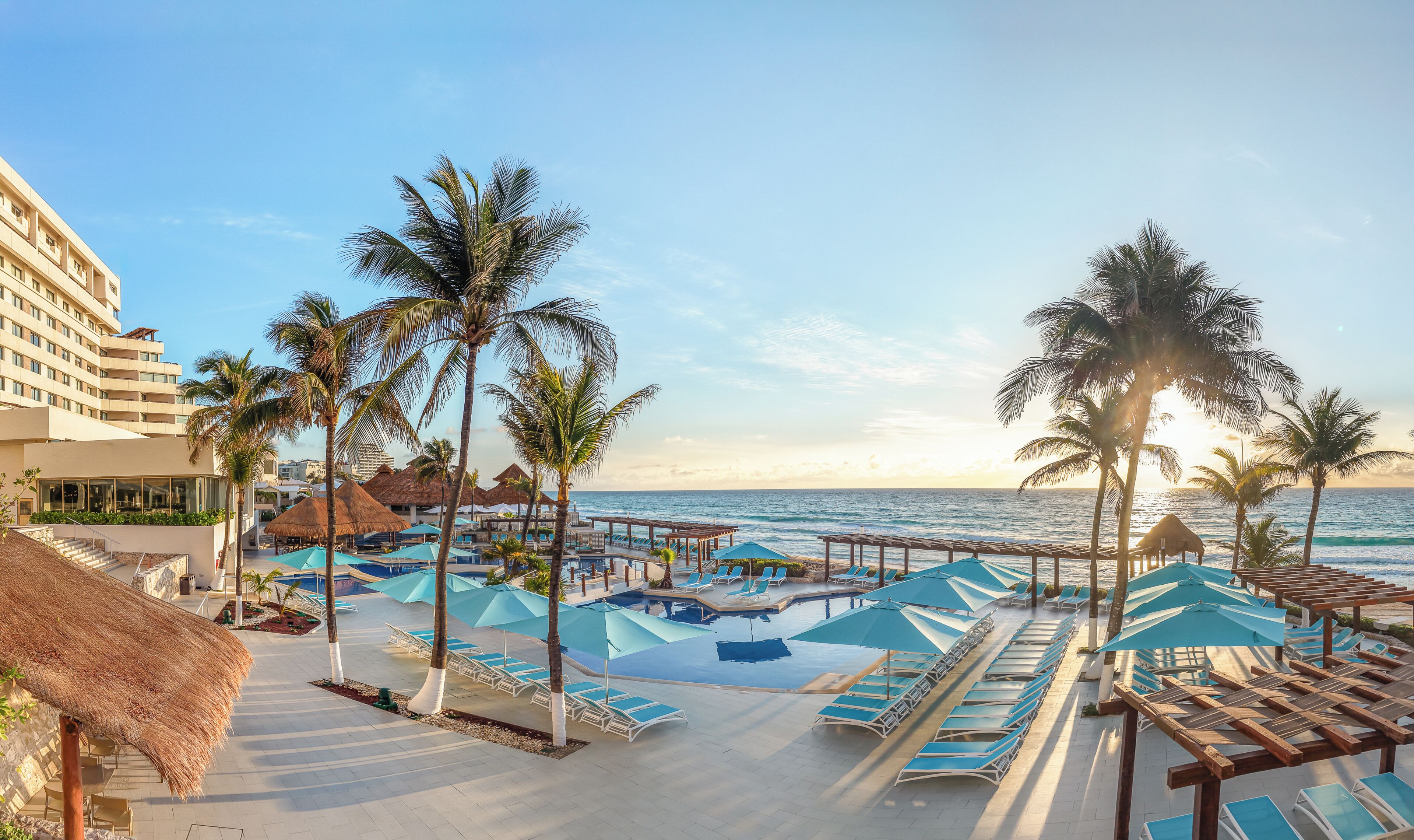 A resort pool area with sun loungers and umbrellas, palm trees, and a view of the ocean.