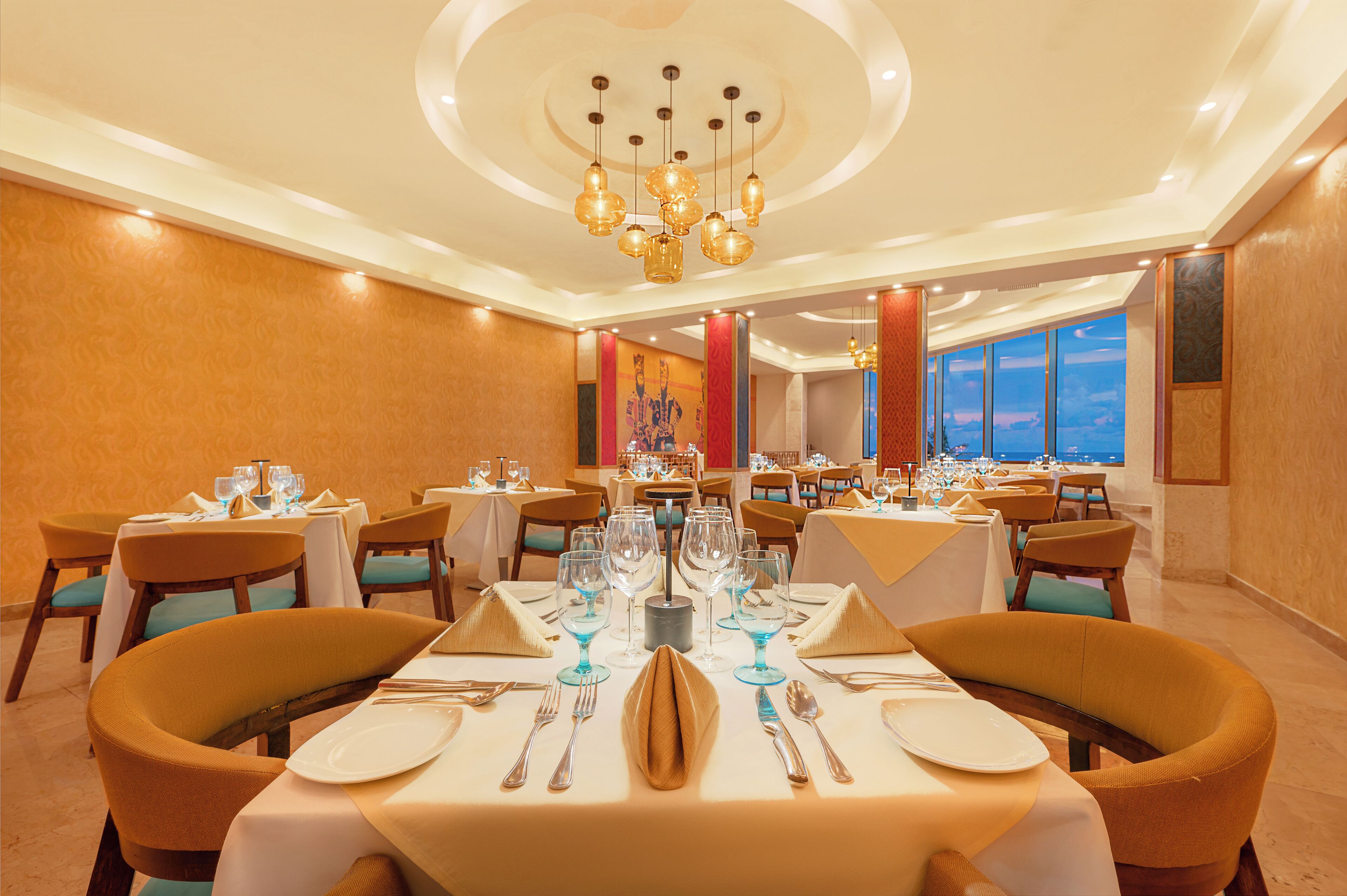 A well-lit dining area with multiple tables set for dining, featuring a central chandelier and a view of the sky through large windows.