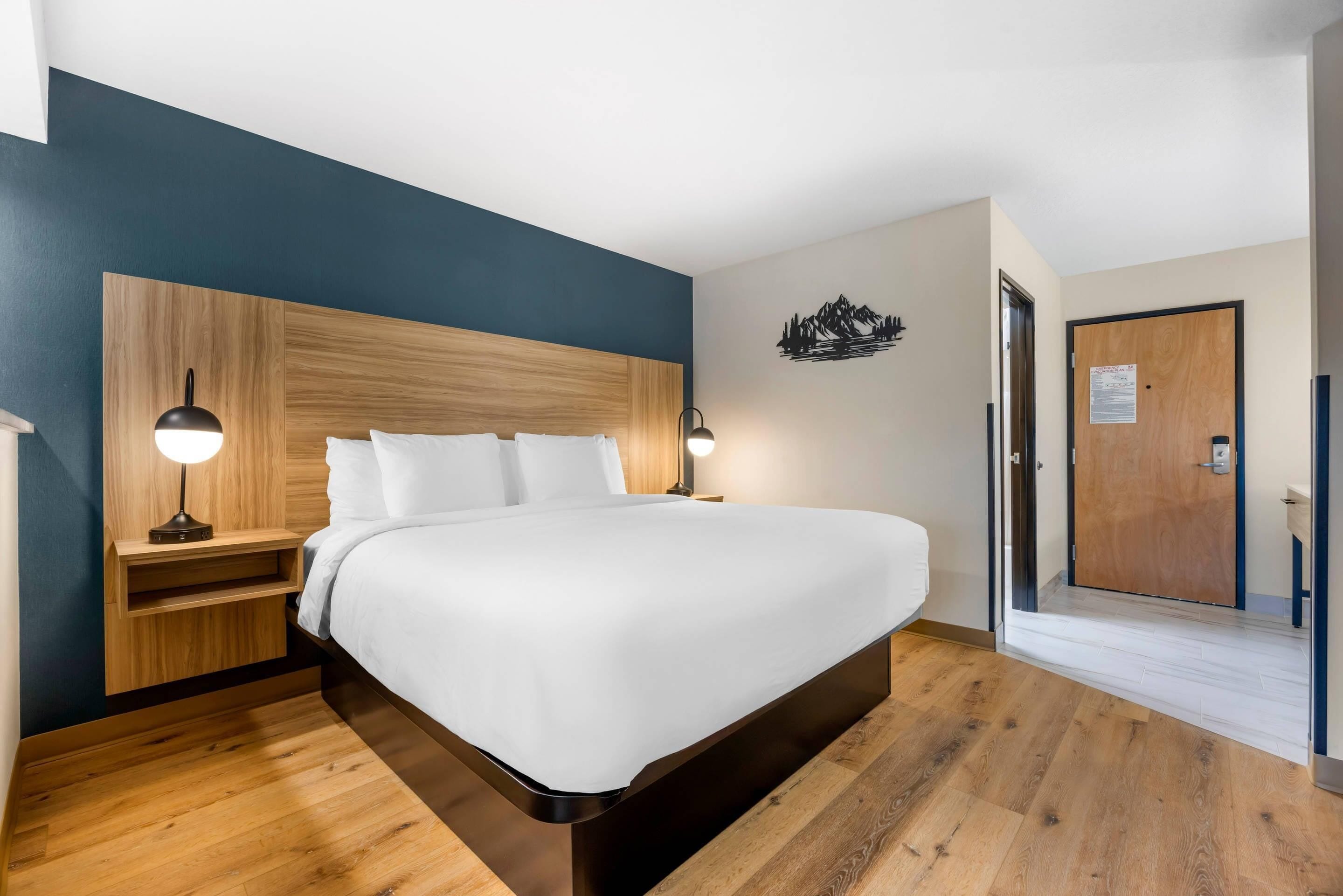 A modern hotel room with a wooden bed frame, white bedding, and a nightstand with a lamp.