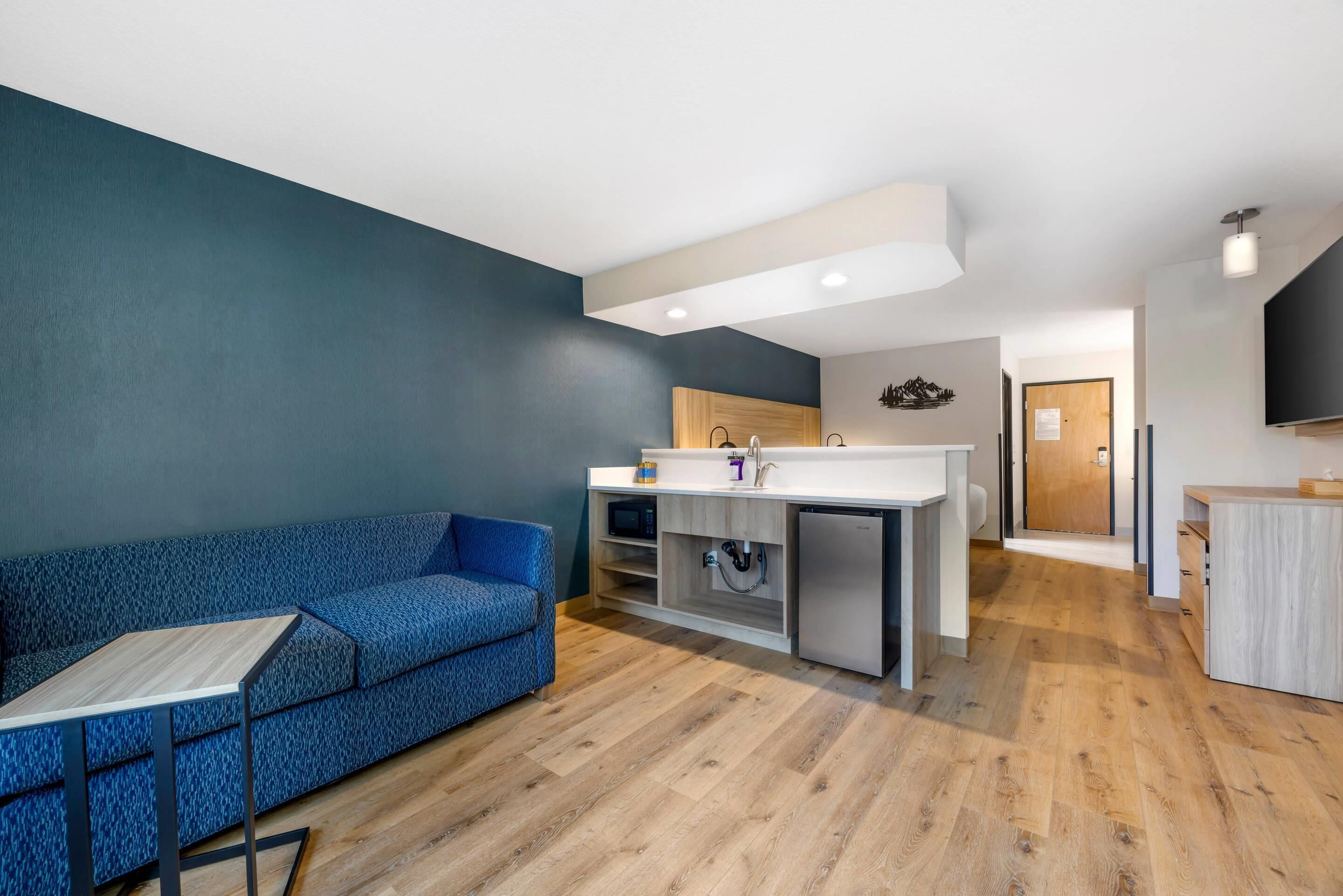 A modern living room with a blue sofa, a small wooden table, and a kitchen area with a sink and cabinets.