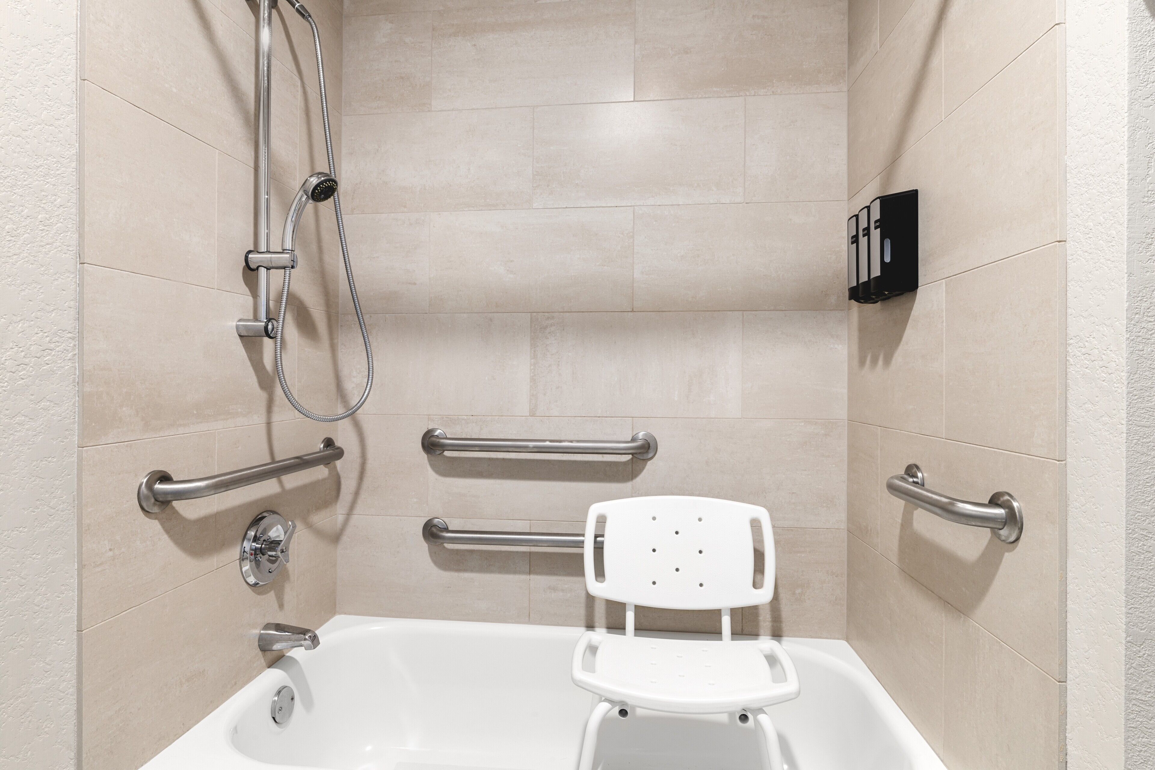 A bathroom with a bathtub, grab bars, a showerhead, and a white chair.