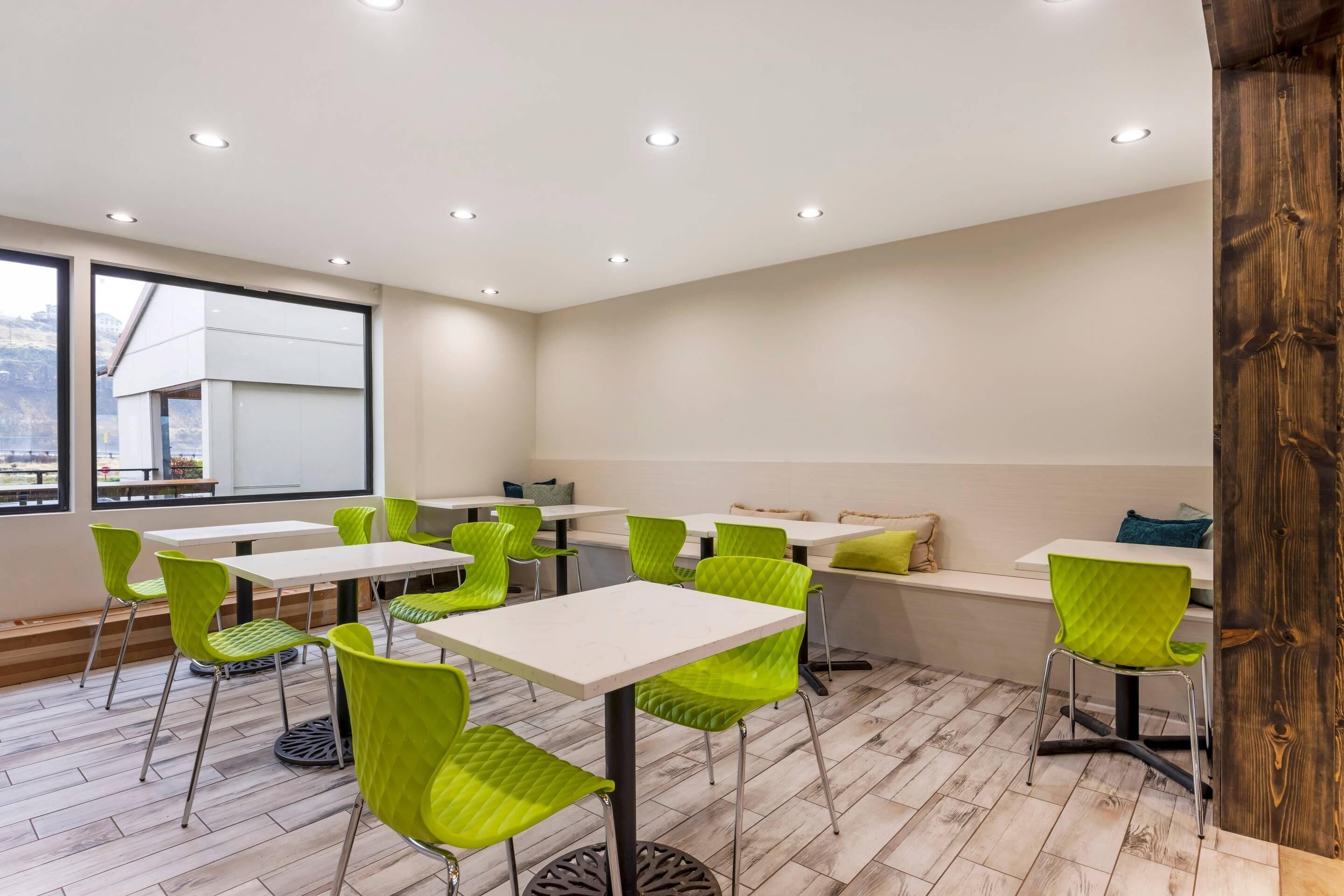 A modern dining area with white tables and green chairs, large windows, and a view of the city.