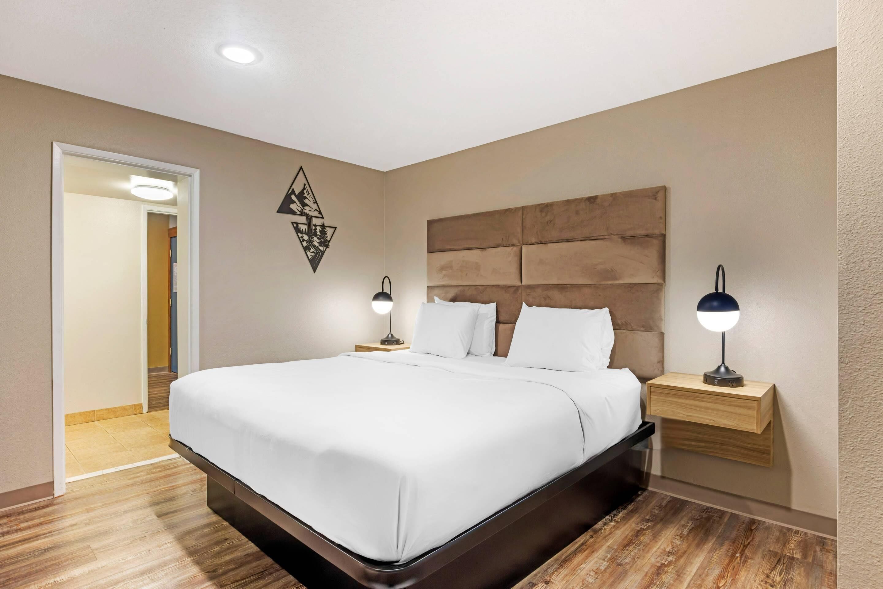 A modern hotel room with a large bed, wooden headboard, bedside tables with lamps, and a geometric wall decoration.