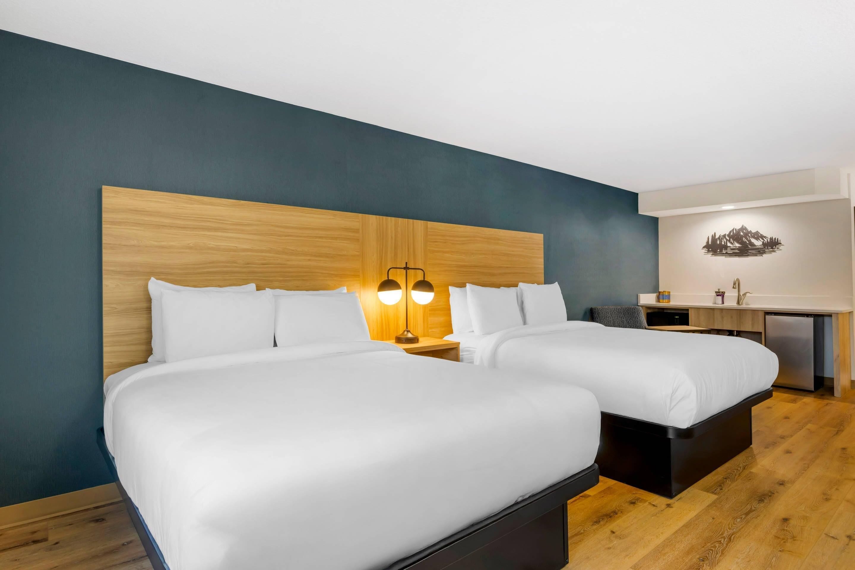 A hotel room with two beds, a wooden headboard, bedside lamps, a small desk, and a wall-mounted artwork.