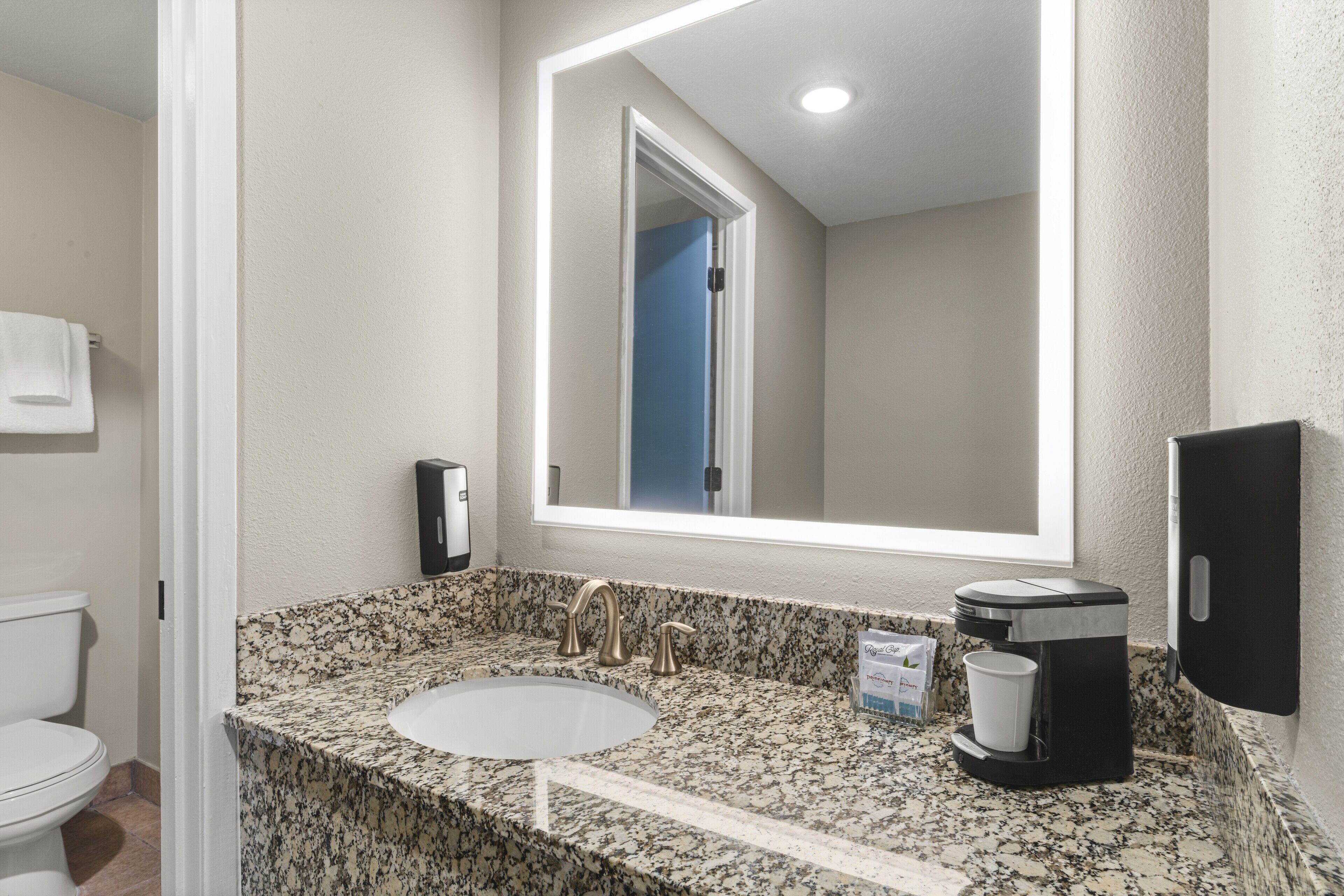 Baño con encimera de granito, lavamanos, espejo, cafetera y secador de manos.