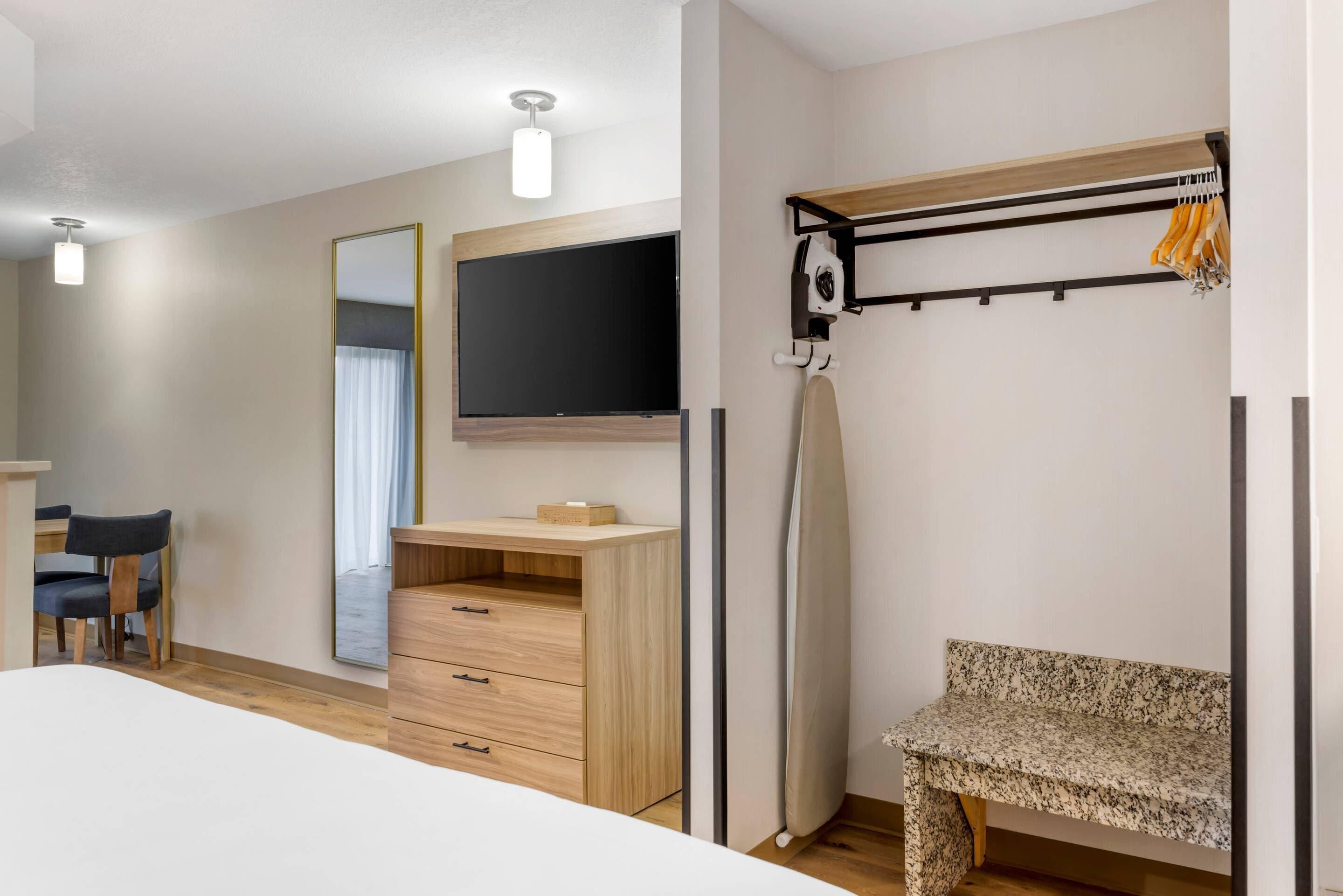 A bedroom with a bed, a desk, a chair, a wardrobe, a mirror, a television, and a bench.