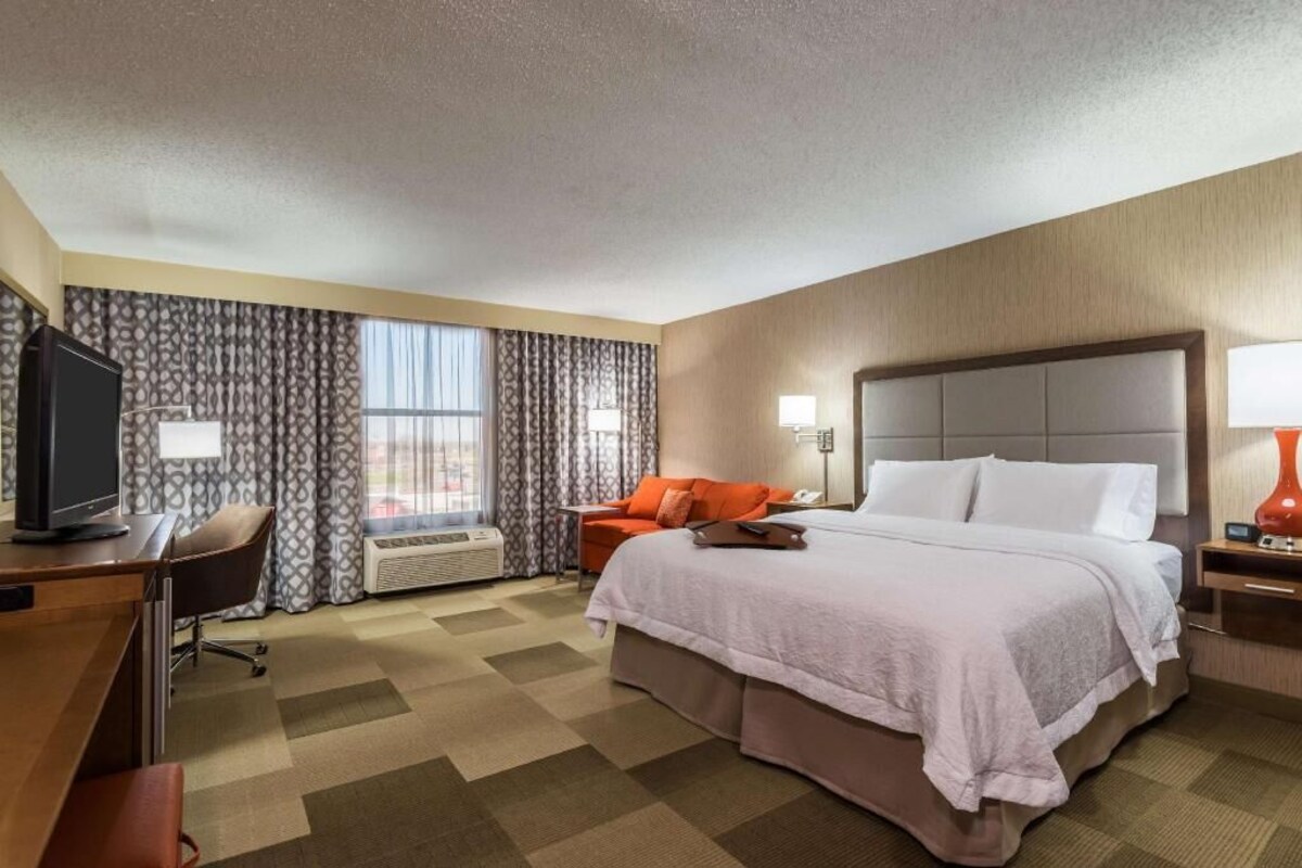 A hotel room with a bed, a desk with a computer, a chair, a TV, a window with curtains, and a bedside table with a lamp.