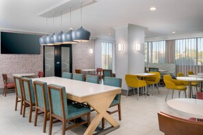 Un espace repas moderne avec une longue table, plusieurs chaises et un téléviseur fixé au mur.