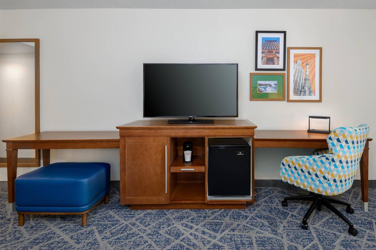 Une chambre d’hôtel avec un bureau en bois, un téléviseur à écran plat, un banc bleu rembourré, une chaise à motifs et des tableaux encadrés accrochés au mur.