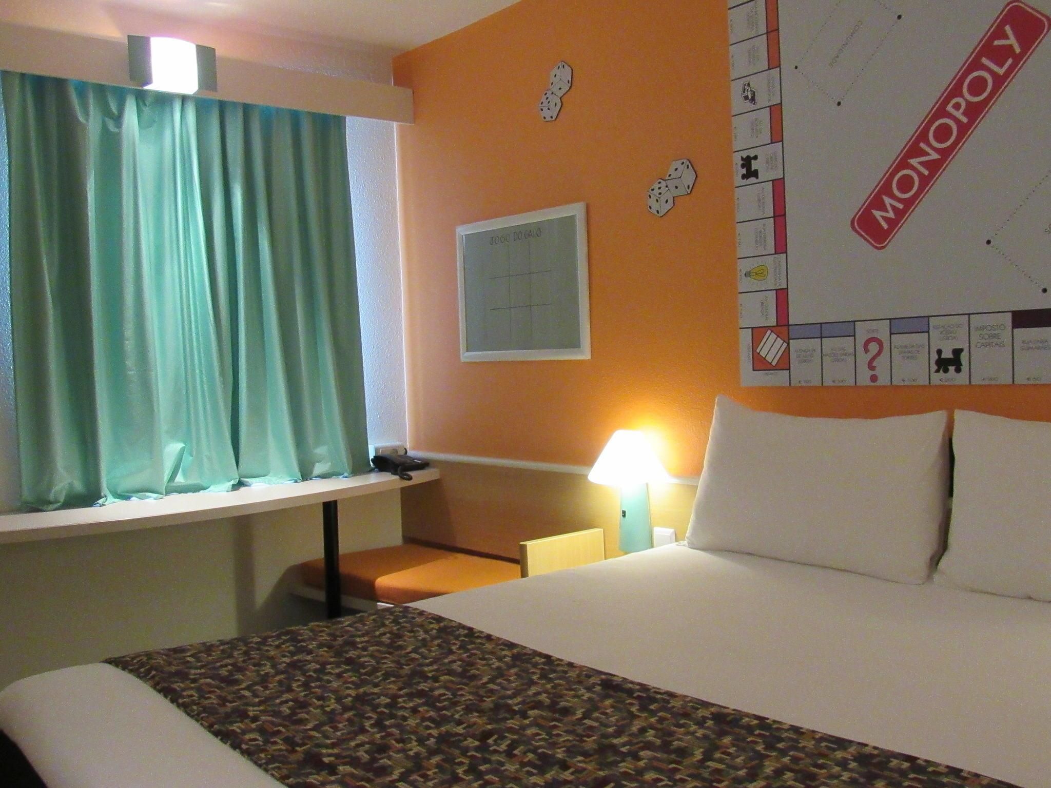 A hotel room with a bed, a desk, a lamp, a Monopoly board game, and a window with curtains.