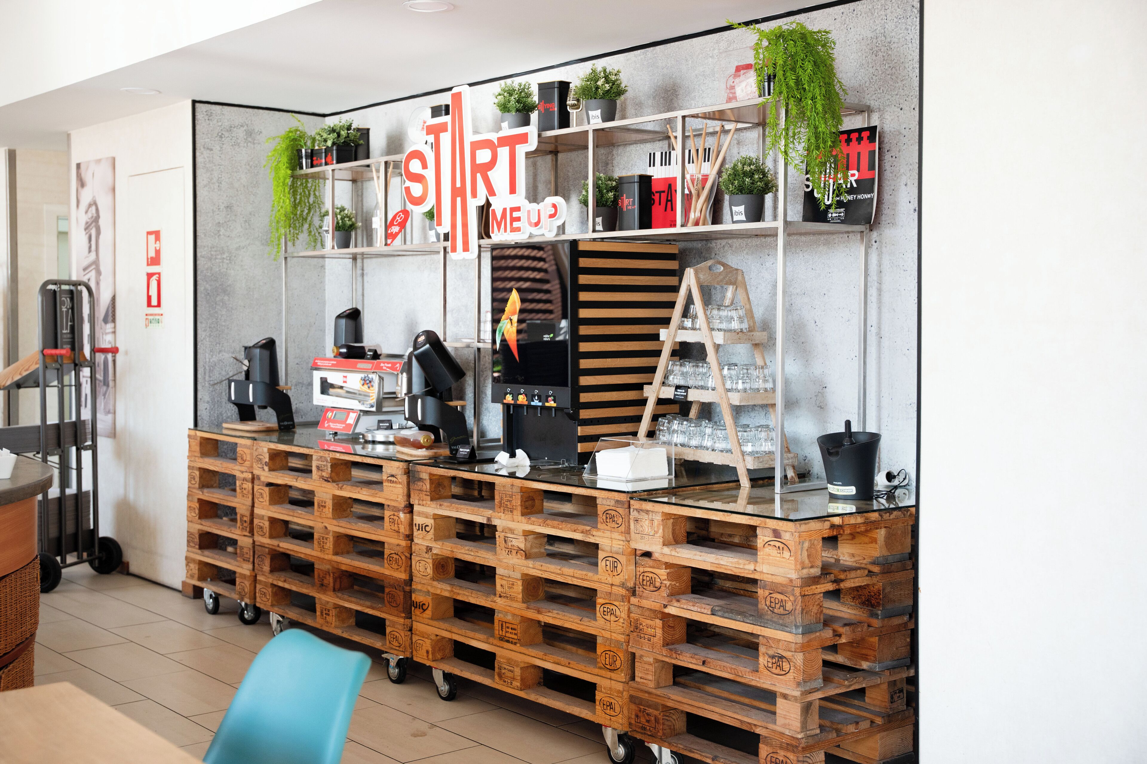 Un espace bar moderne avec des caisses en bois servant d’étagères, une machine à café et un panneau sur lequel on peut lire : « START ME UP ».