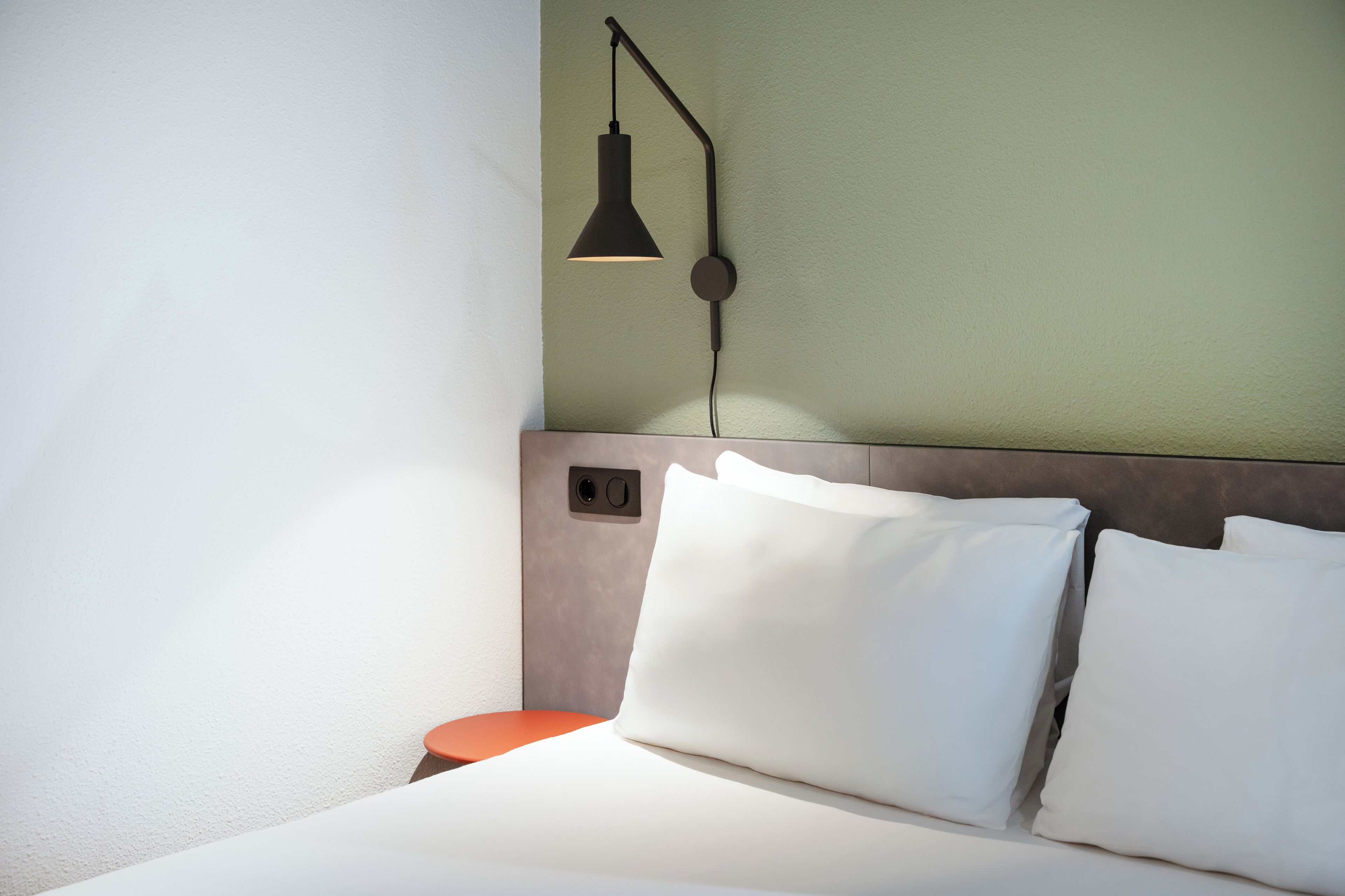 Een net opgemaakt bed met witte lakens en kussens, een klein nachtkastje in oranje en een wandlamp.