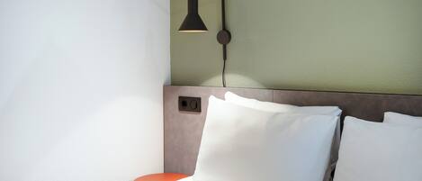 Een net opgemaakt bed met witte lakens en kussens, een klein nachtkastje in oranje en een wandlamp.