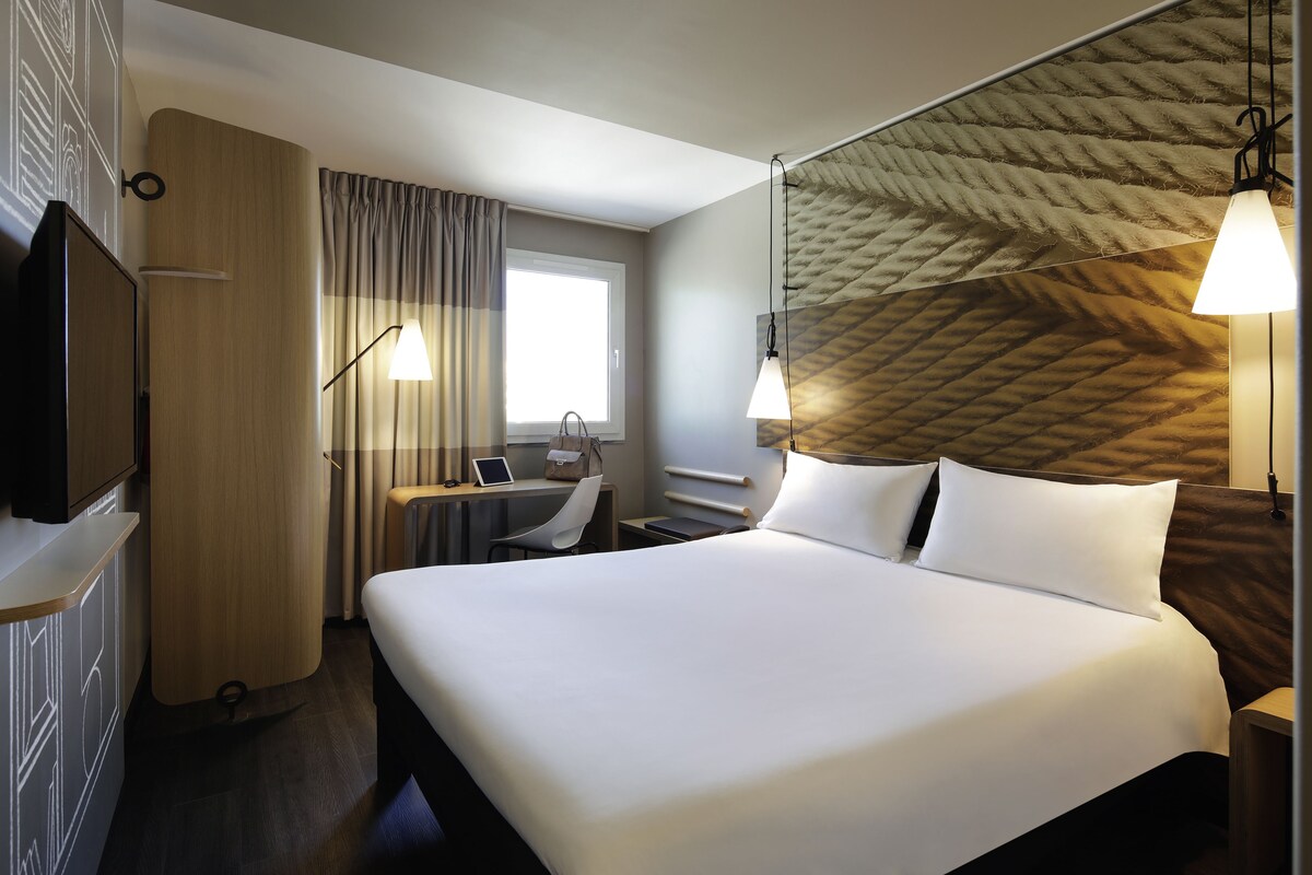 A hotel room with a large bed, a desk with a laptop, a TV, and a shower area.