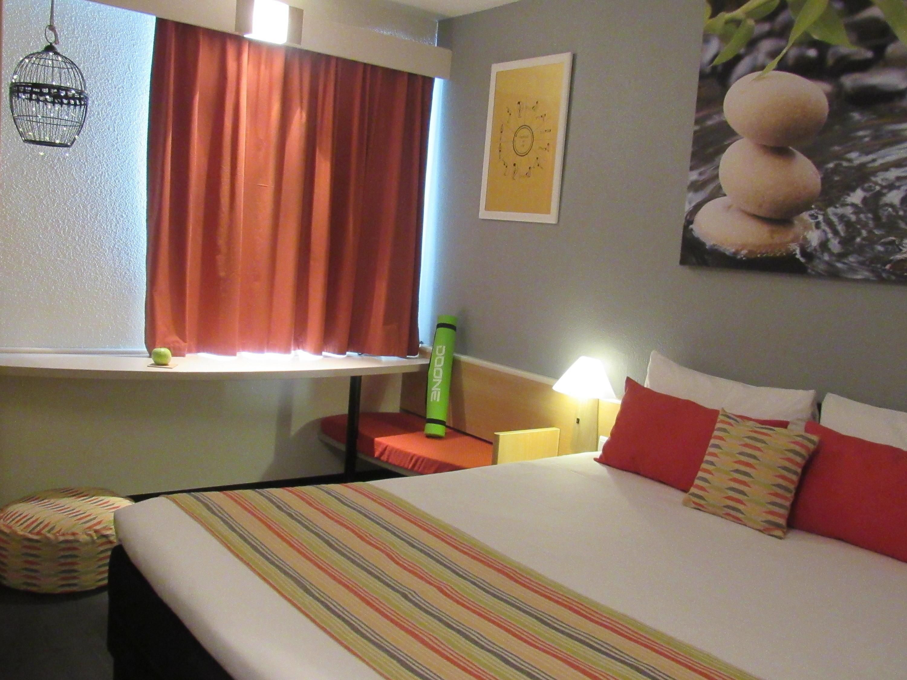 Habitación de hotel con cama, ventana con cortinas, lámpara, reloj de pared y una pequeña planta.