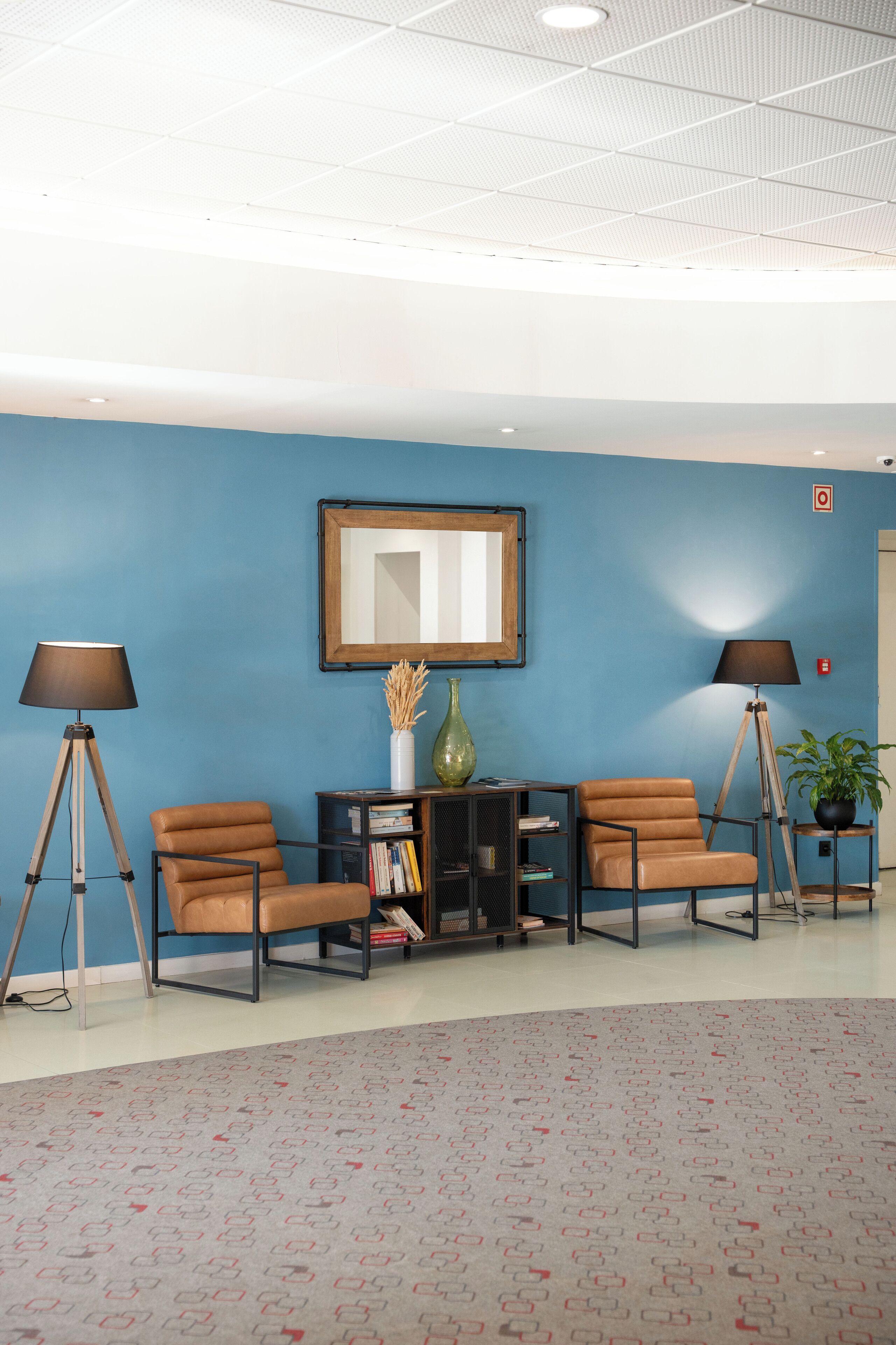 A reception area with blue walls, two brown leather chairs, a wooden side table, a floor lamp, a bookshelf, and a mirror.