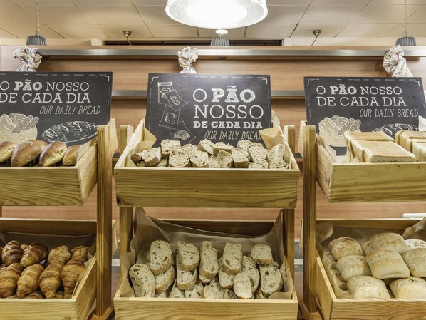 A bakery display with various breads and pastries in wooden crates, labeled 'O PÃO NOSSO DE CADA DIA OUR DAILY BREAD'.
