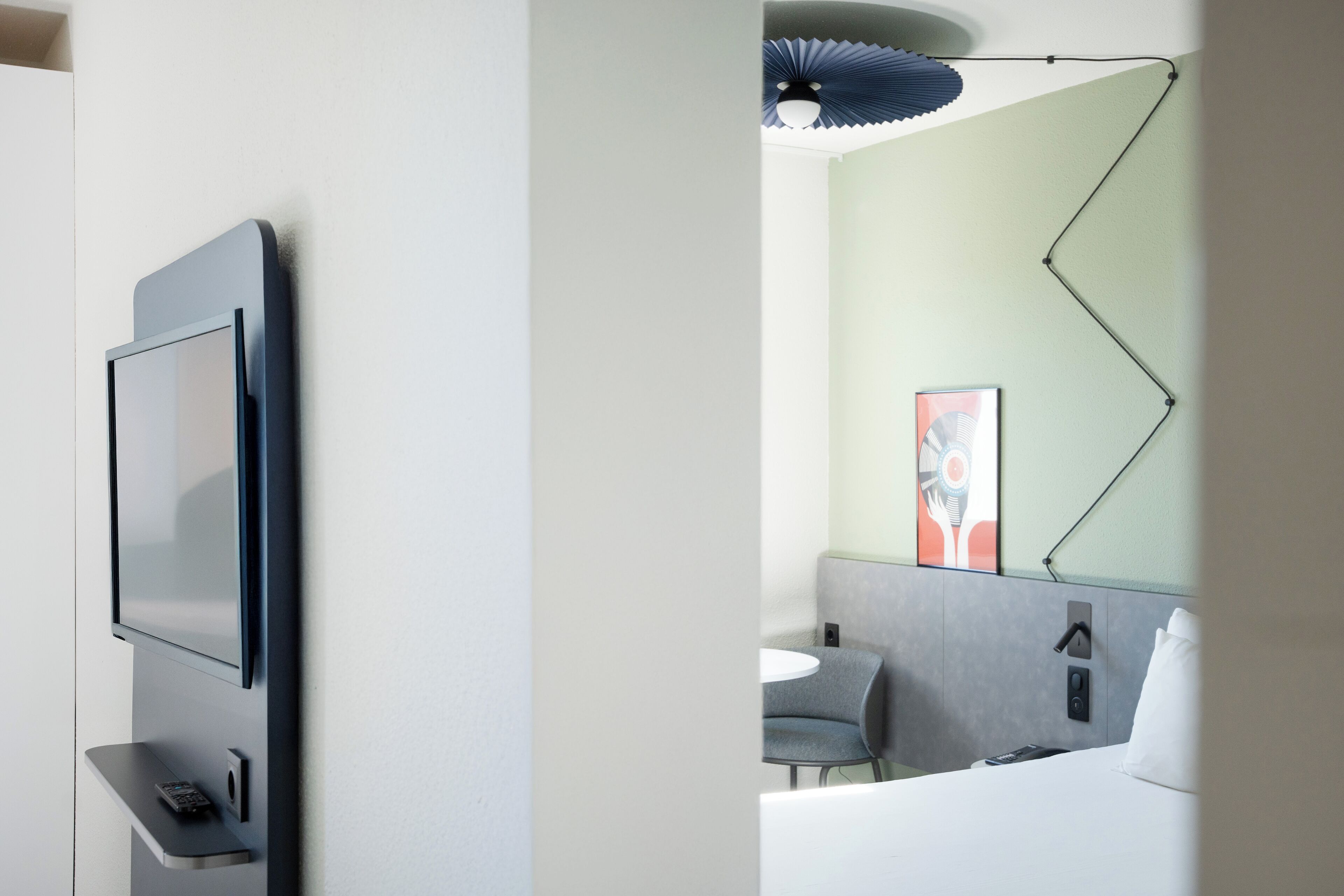 Habitación de hotel moderna con una televisión de pantalla plana en la pared, un escritorio pequeño con un teléfono, una cama con cabecera blanca, un sofá gris y una lámpara de techo original.